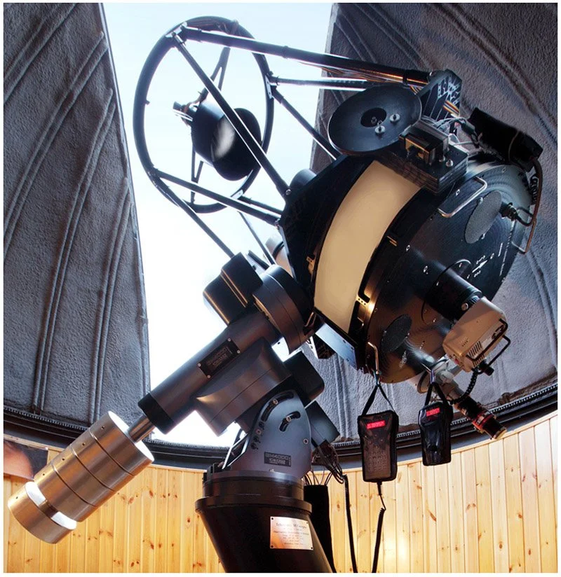 A large telescope with a white protective cover on the main tube, mounted inside an observatory with wooden walls and a circular ceiling, pointing upwards towards the sky.