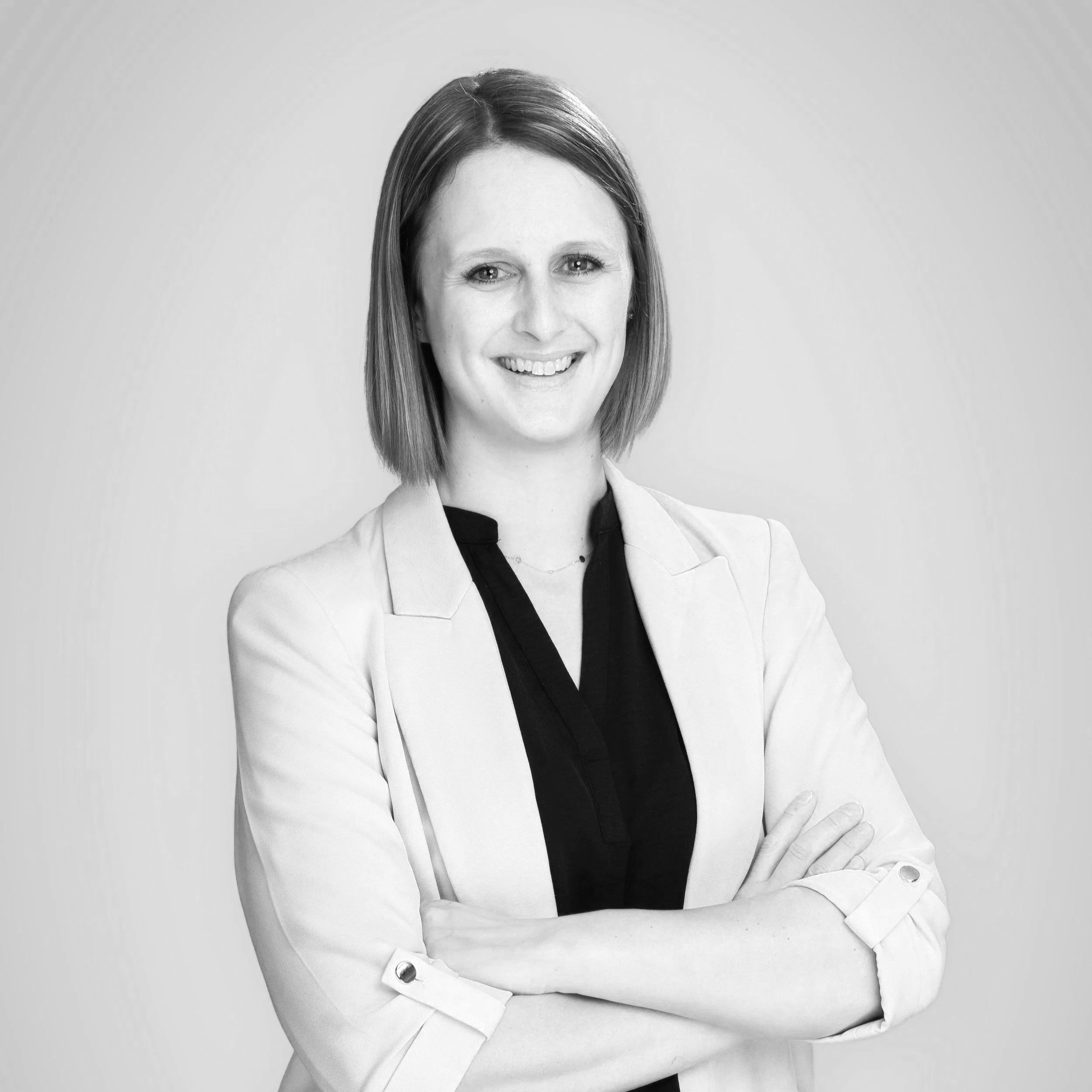 Schwarz-weiß Porträt einer lächelnden Frau mit kurzen, glatten Haaren, in einem hellen Blazer und schwarzem Top, vor hellem Hintergrund, mit verschränkten Armen.