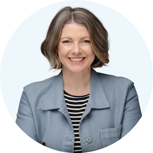 Eine Frau mit kurzem braunem Haar, die lächelnd in die Kamera schaut, trägt eine blaue Jacke und ein schwarzes und weiß gestreiftes Oberteil.