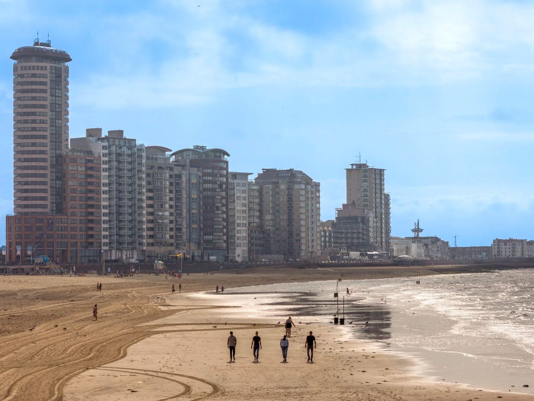 View of a city skyline with tall buildings along a beach. Few people walk on the sandy shore, and the tide is low with wet sand reflecting the sky.