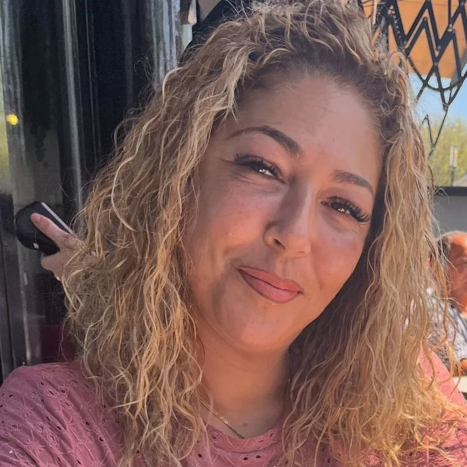 A woman with curly red hair sitting on a couch, resting her chin on her hand, smiling gently at the camera.