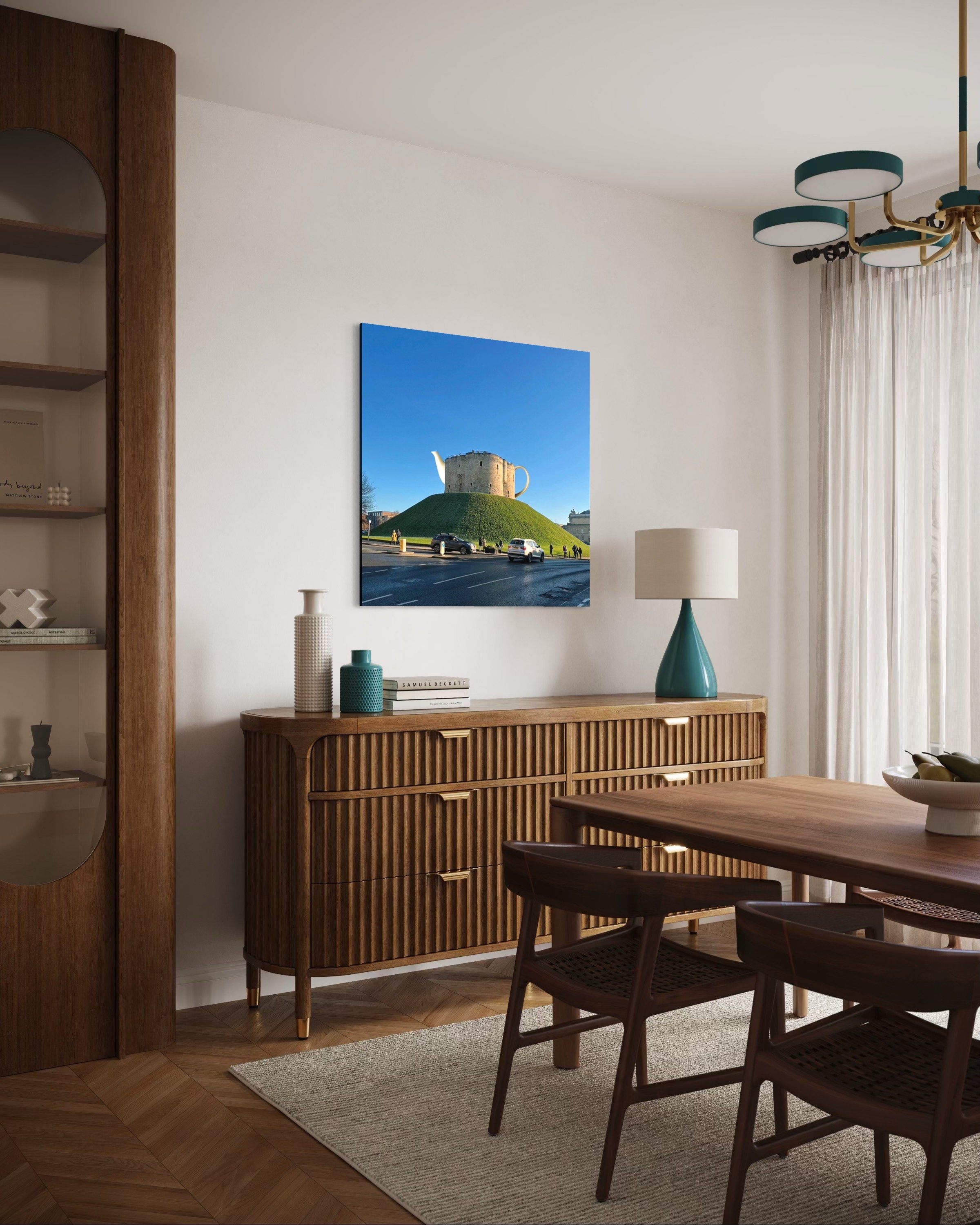 A living room with a wooden sideboard and a large blue teapot-shaped lamp, a white textured vase, two blue vases, and a stack of books on top. Above, a framed photograph of a castle on a grassy hill under a blue sky. To the right, a window with sheer