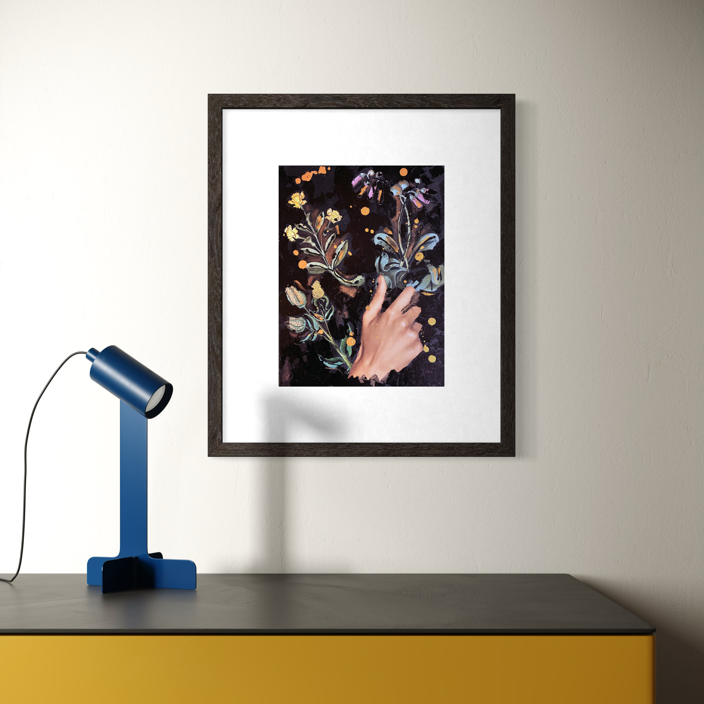 Framed artwork depicting a hand touching swirling floral patterns with a dark background and colorful accents, placed on a wall above a dark-colored surface with a blue desk lamp.