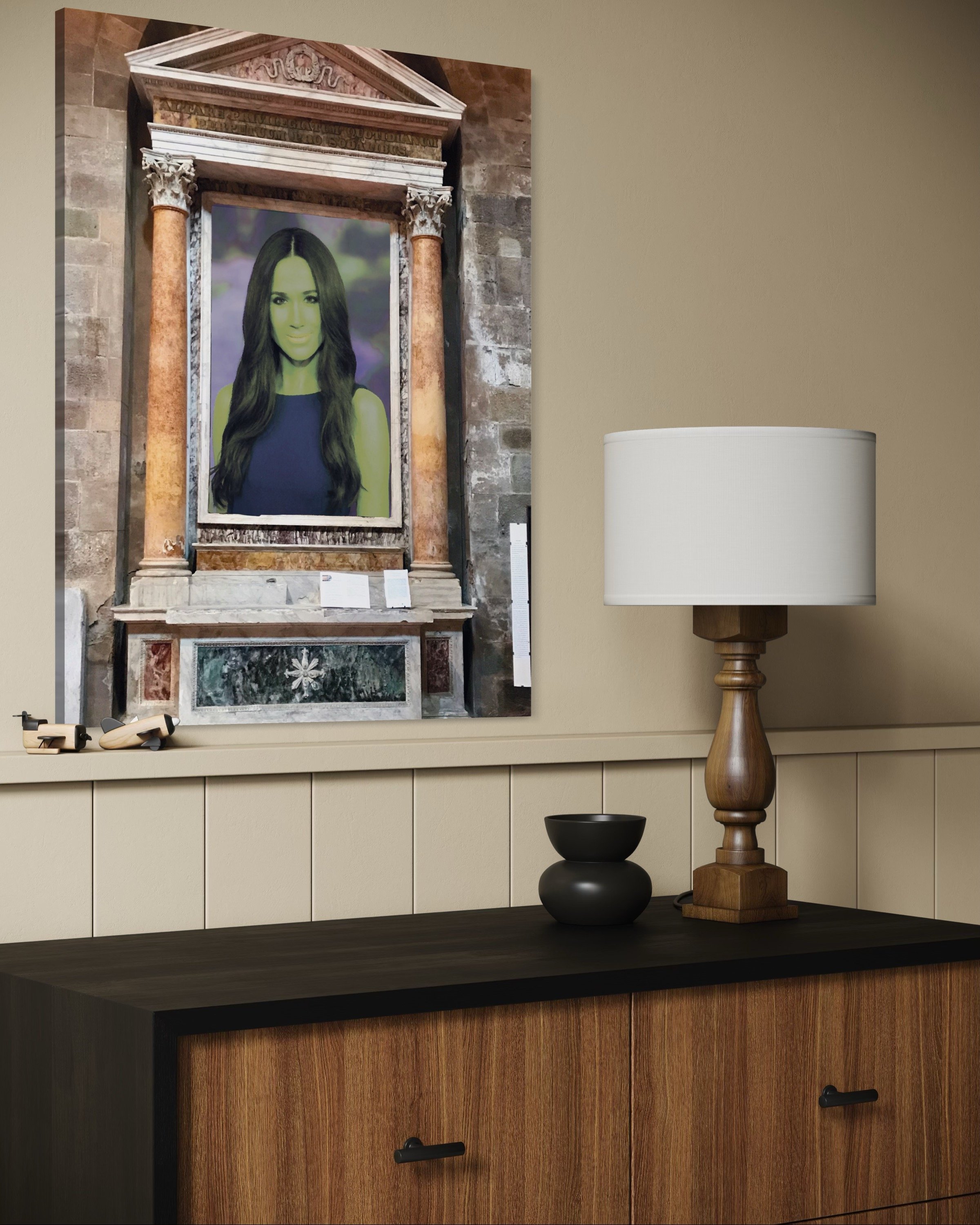 A portrait of a woman with long dark hair on a wall behind a wooden sideboard, with a lamp and black decorative object on the sideboard in a room with beige walls.