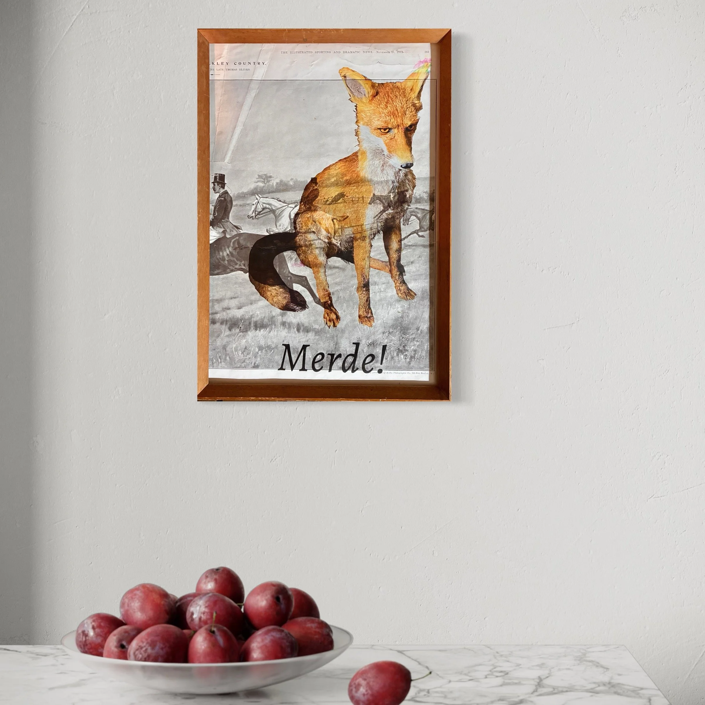 Framed vintage poster of a fox with a background of horses and a rider, and the word "Merde!" at the bottom, hanging on a plain white wall.