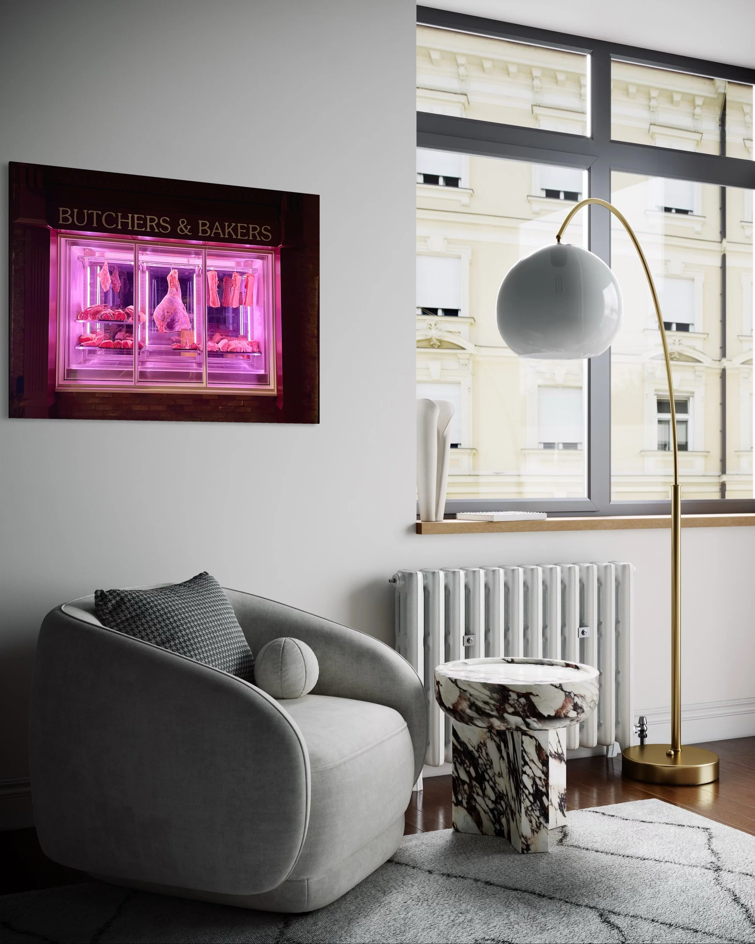 A modern living room with a gray armchair, a marble side table, a white radiator, a tall white and gold floor lamp, and a large window showing an exterior view of a building. A pink-lit display case labeled "Butchers & Bakers" with meat and baked goo