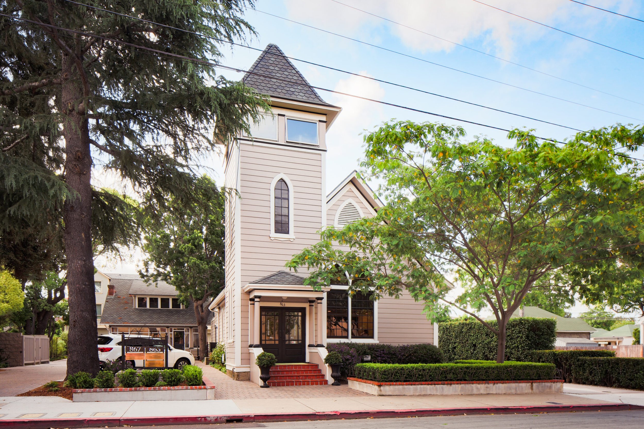A beige Victorian-style house with a steep roof, Gothic windows, located on a street with trees and manicured bushes.