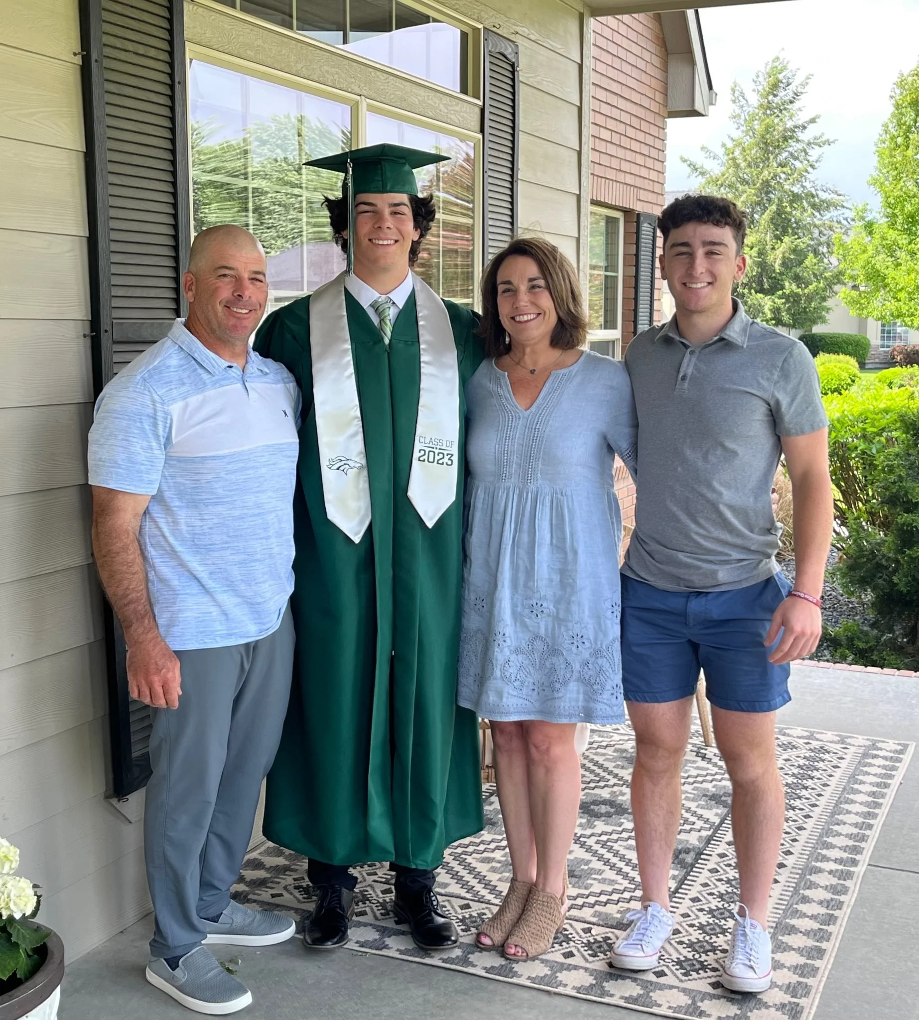 Family standing on porch with graduate in cap and gown, smiling, during daytime.
