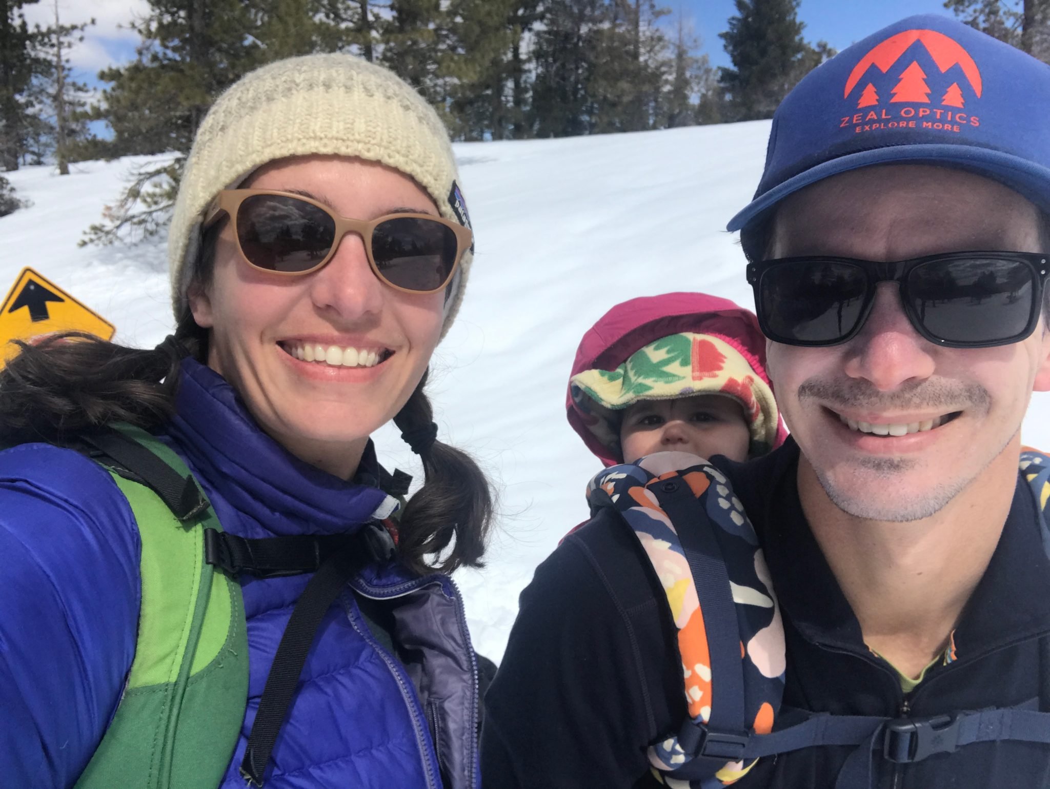 Family taking a selfie in a snowy mountain landscape, two adults wearing sunglasses and a child with a colorful hat strapped to a man's backpack.