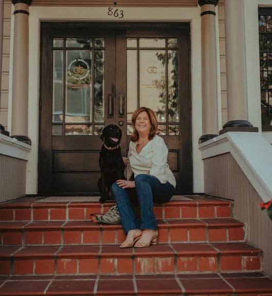 A woman with brown hair sitting on brick steps in front of a house door, next to a black dog, both smiling.