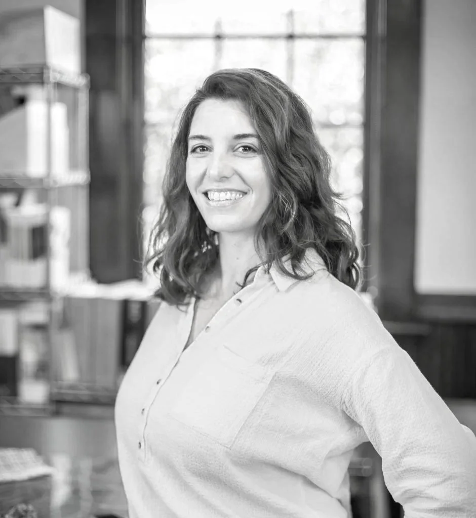 Black and white photo of a woman with curly hair smiling in an indoor setting, possibly a kitchen or café.