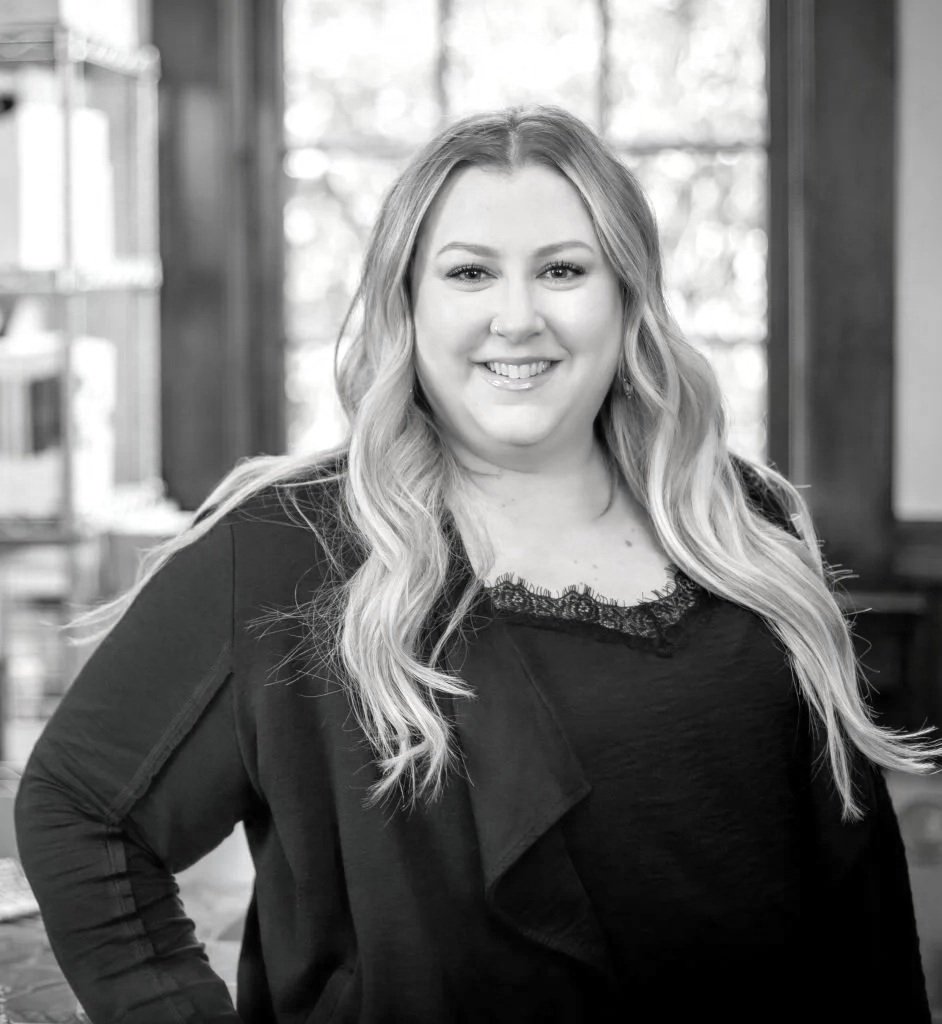 A smiling woman with long wavy hair, wearing a dark top with lace detail and a blazer, standing indoors with large windows in the background.