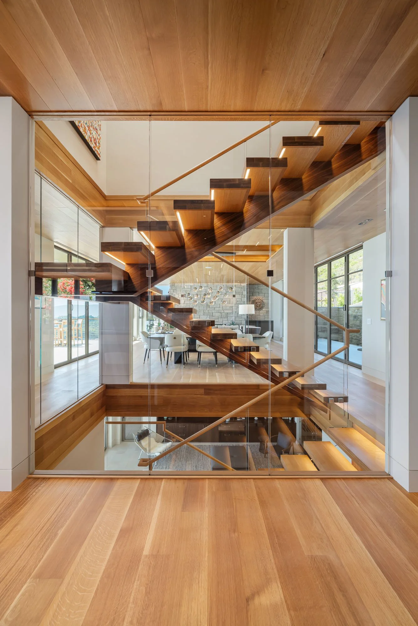Interior view of a modern house showcasing a floating wooden staircase with glass walls, overlooking a dining area and living room with large windows.