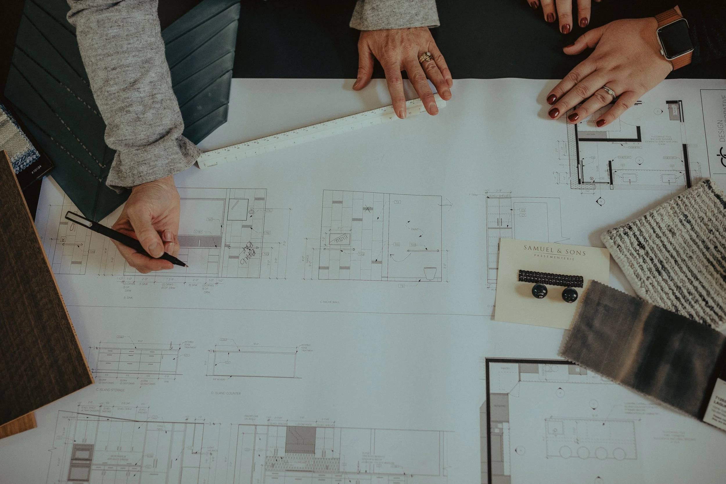 People examining architectural blueprints with color samples and writing tools on a desk.
