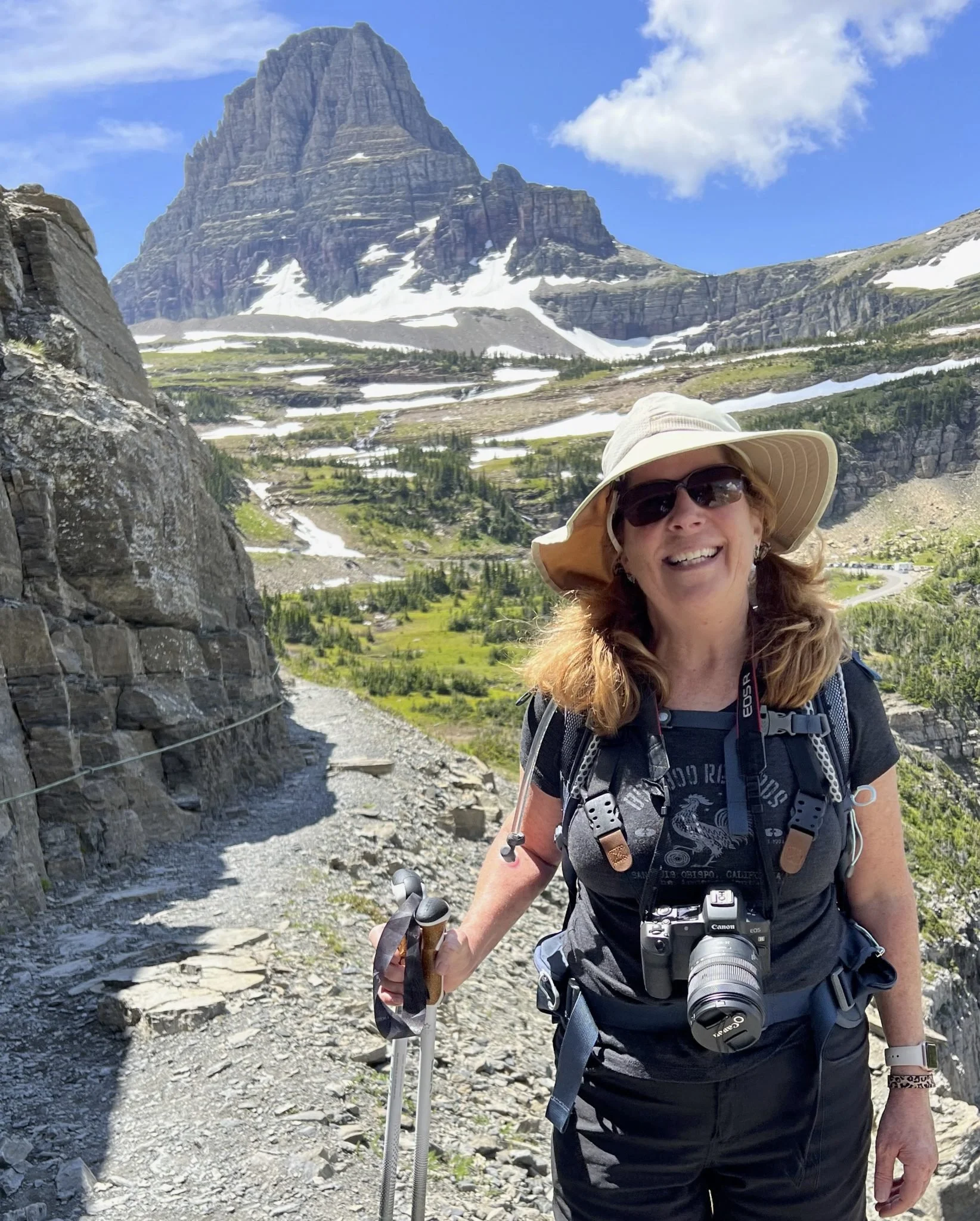 Woman hiking on a mountain trail with a camera around her neck, wearing a wide-brimmed hat and sunglasses, with snow-capped mountain and green valley in the background.