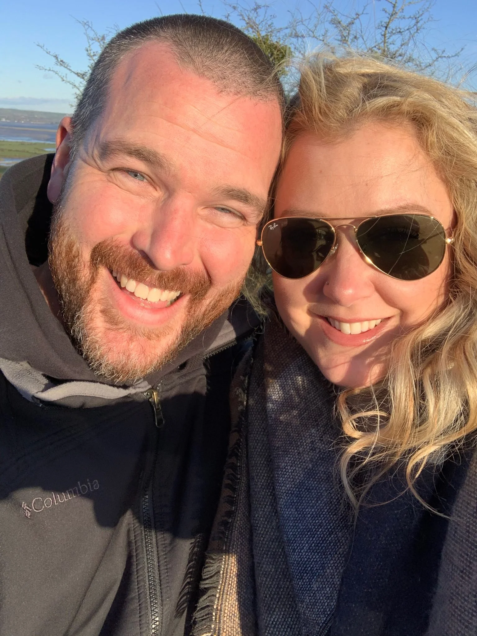 A smiling man with a beard and a woman wearing sunglasses taking a close-up selfie outdoors with a landscape and blue sky in the background.