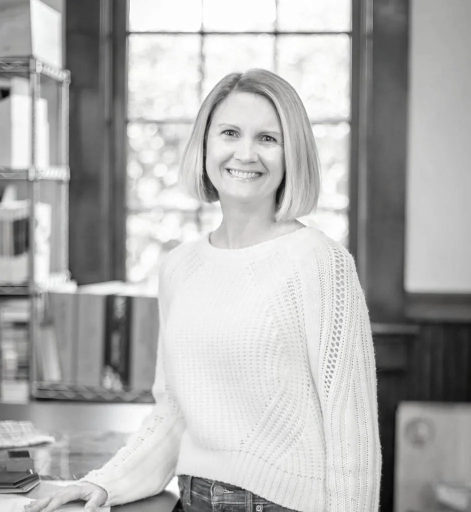 A smiling woman with shoulder-length hair in a bright room with large windows, wearing a white knitted sweater.