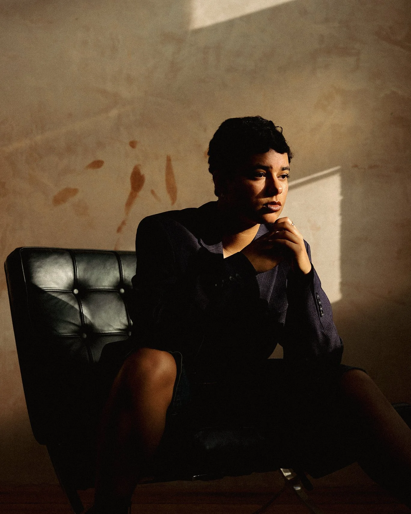 A woman sitting on a black leather chair, with her hands clasped near her chin, looking thoughtful in a dimly lit room with sunlight casting shadows on the wall behind her.