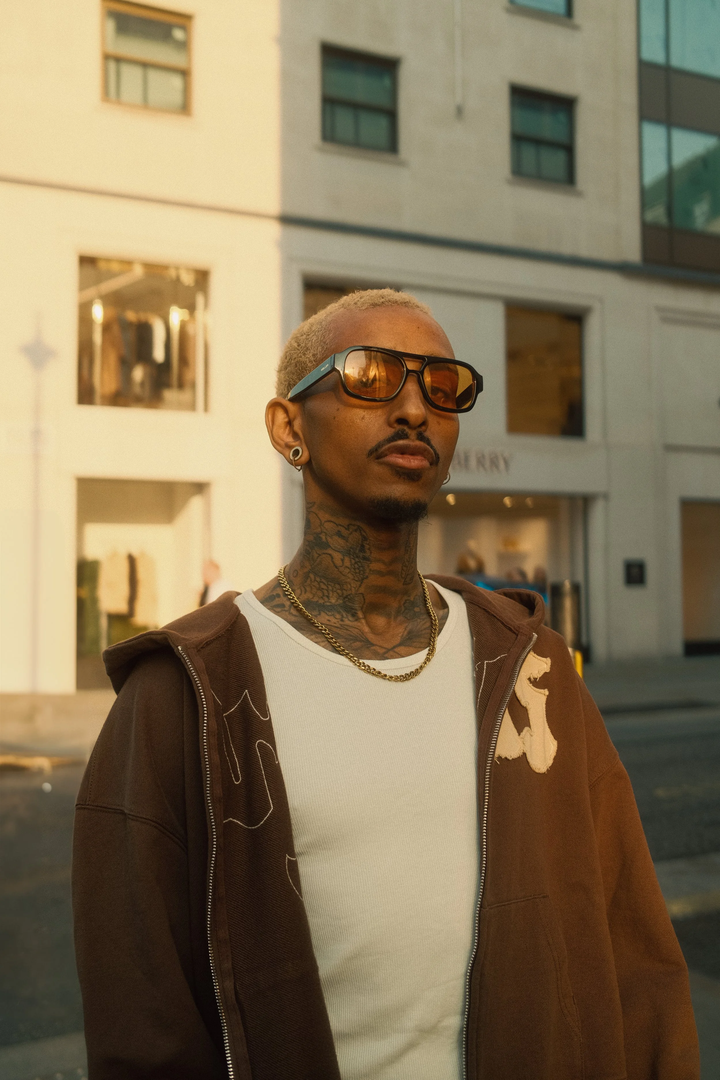 A man with tattoos and blonde hair wearing sunglasses, a white shirt, and a brown hoodie standing outdoors in an urban area during sunset.