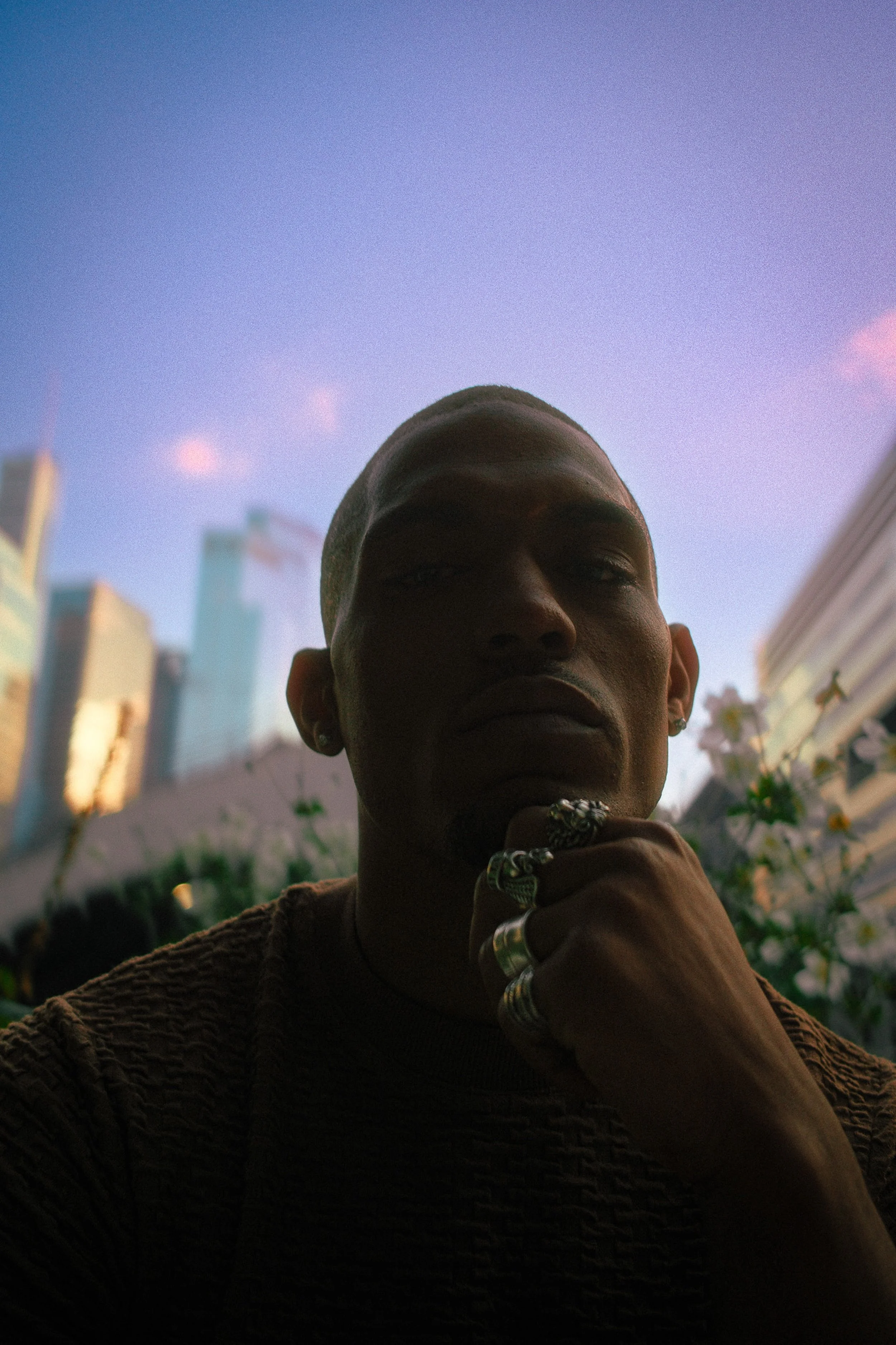 A person with dark skin, wearing multiple rings and earrings, is posing outdoors against a cityscape background with tall buildings and a purple sky.