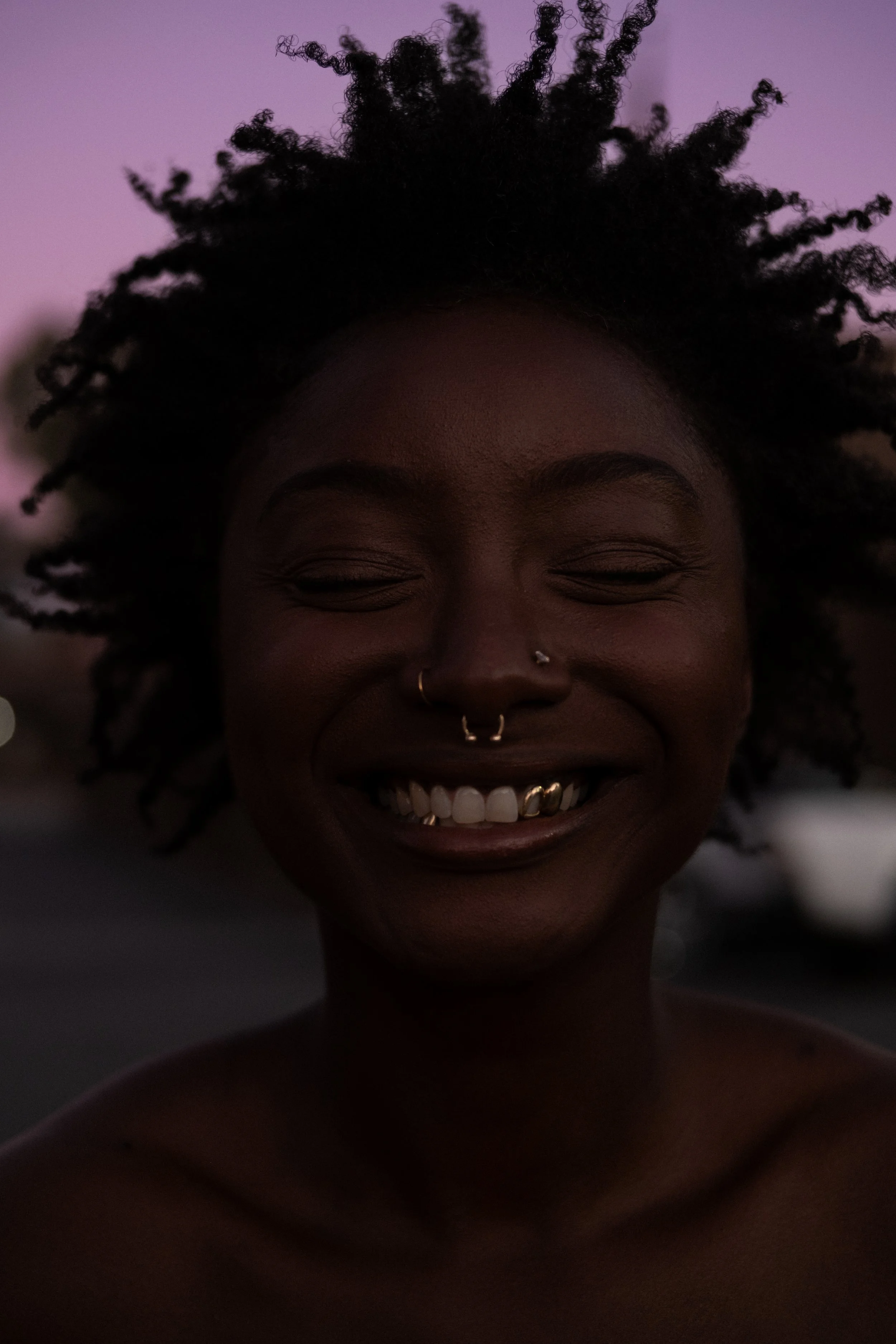 A woman with dark skin and curly hair smiling with closed eyes during sunset, showing her gold teeth and multiple nose piercings.