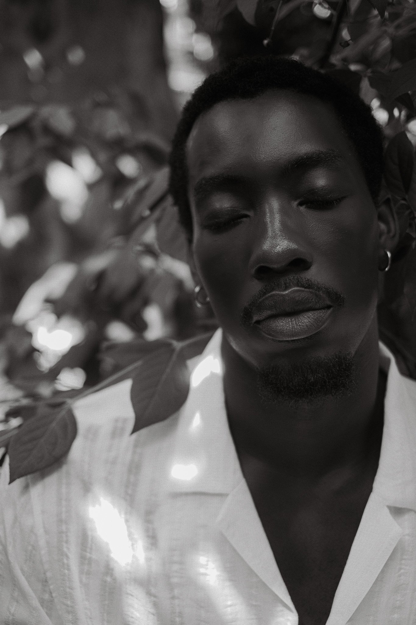 A black and white close-up photo of a person with short hair, earrings, a mustache, a beard, and a white shirt, with leaves in the background.