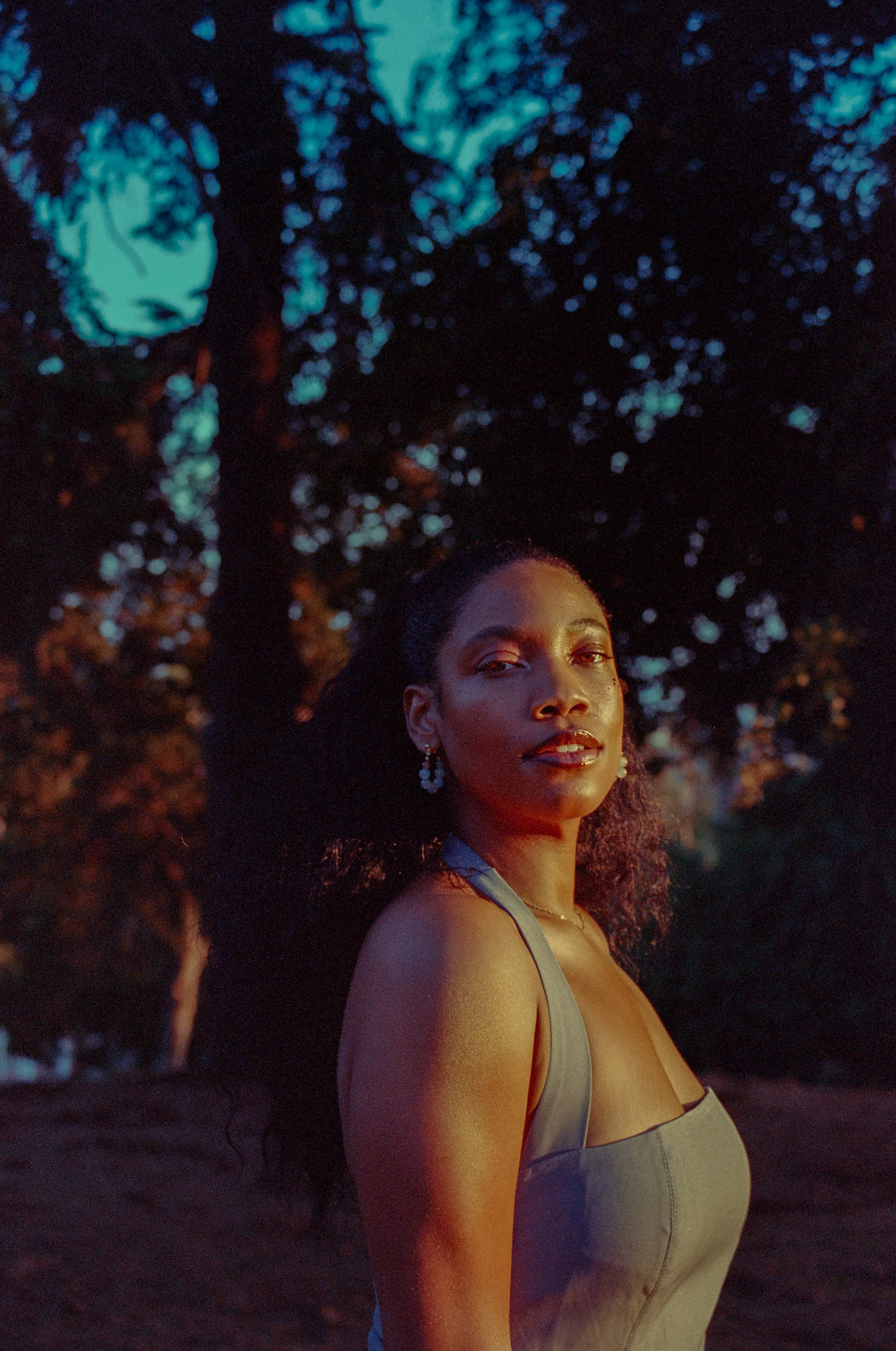 A woman standing outdoors at sunset, looking at the camera, with trees in the background.