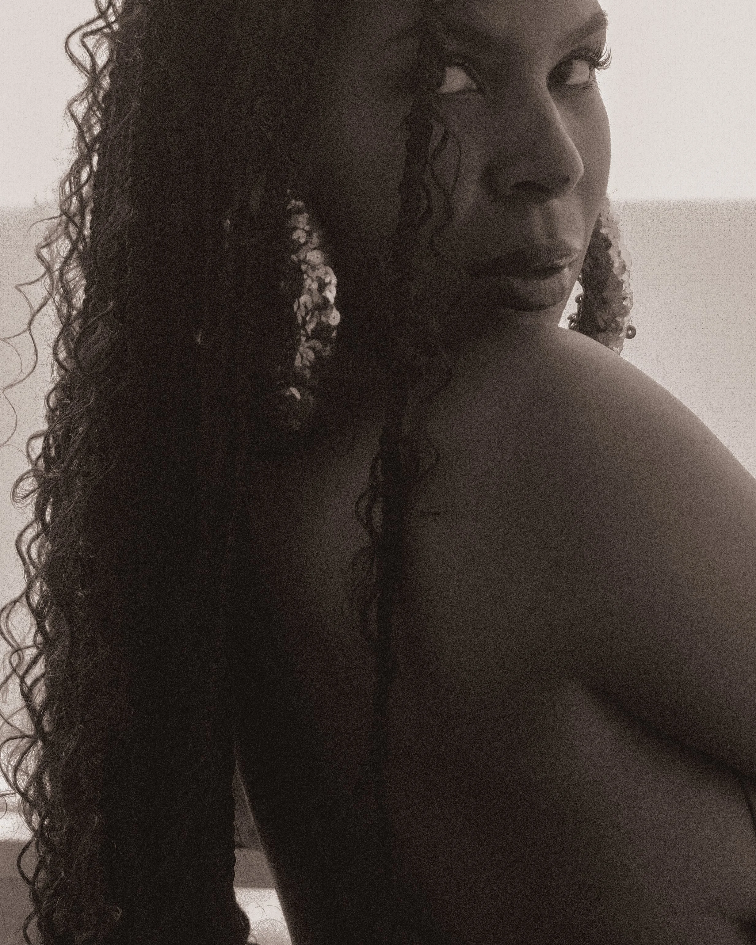 A black and white close-up photo of a woman with curly hair, earrings, and makeup. She is looking over her shoulder with a serious expression.