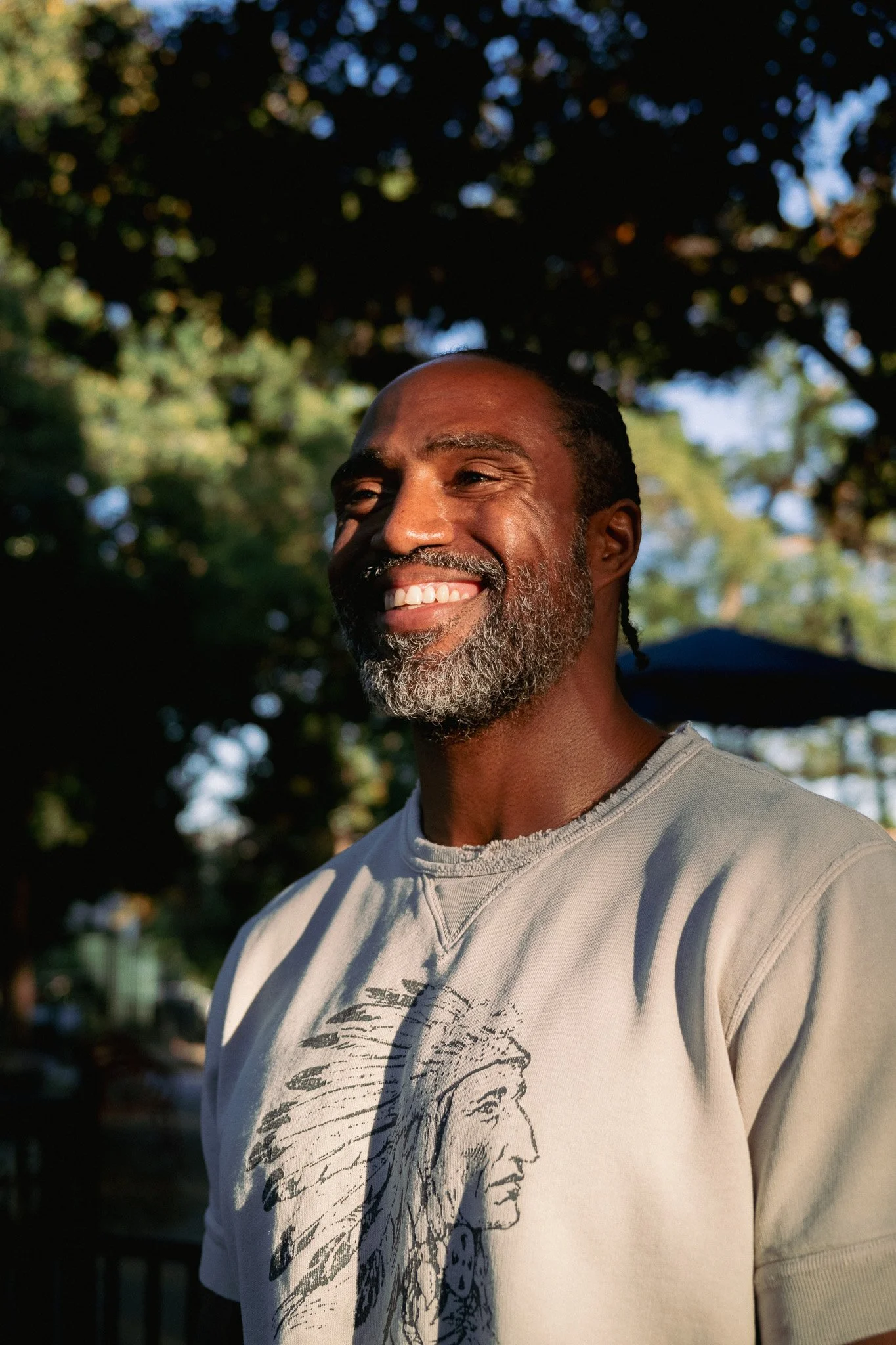 A smiling African American man with a beard and dreadlocks standing outdoors in a park during sunset, with trees and a dark umbrella in the background.