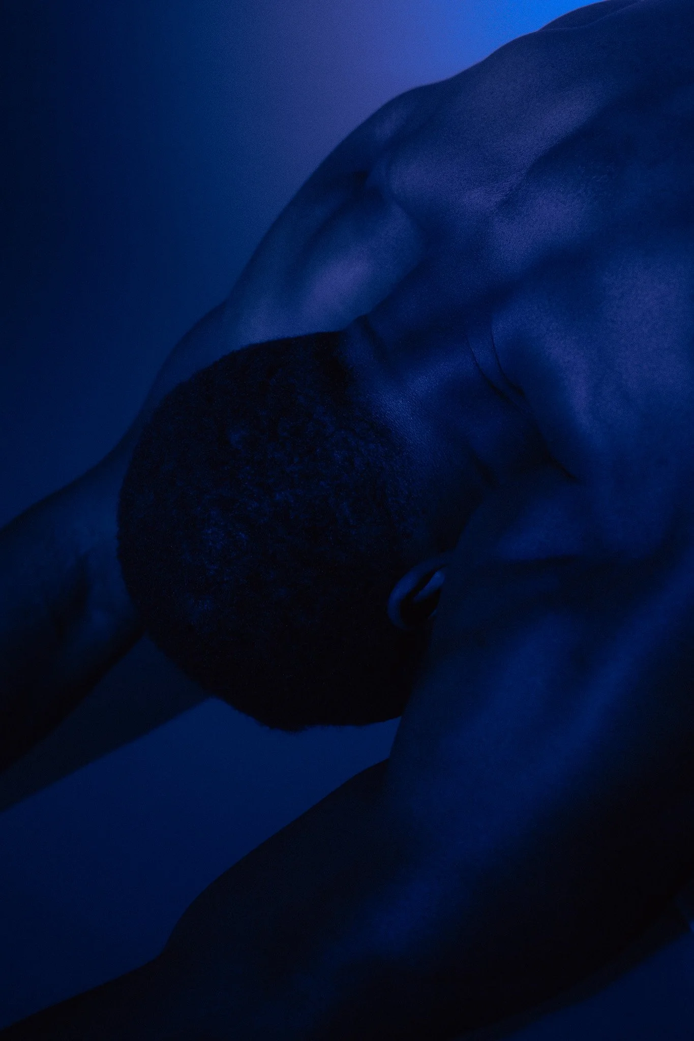 Close-up of a person with dark curly hair and a hoop earring, resting their head on their arm in bluish lighting.