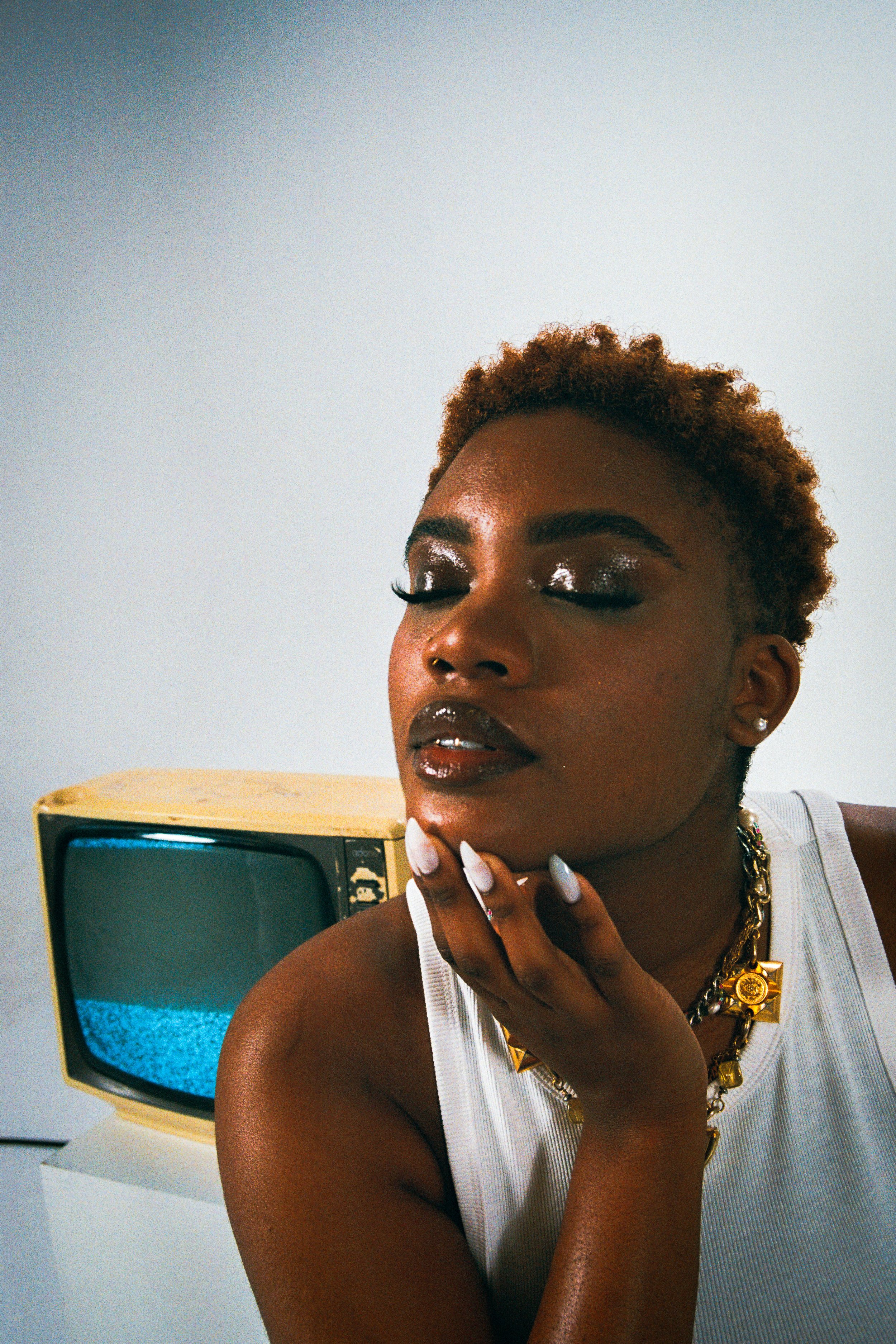 A woman with short curly hair and dark makeup with glittery eyeshadow and glossy lips, wearing jewelry, touching her face with her hand, in front of a vintage TV showing static.