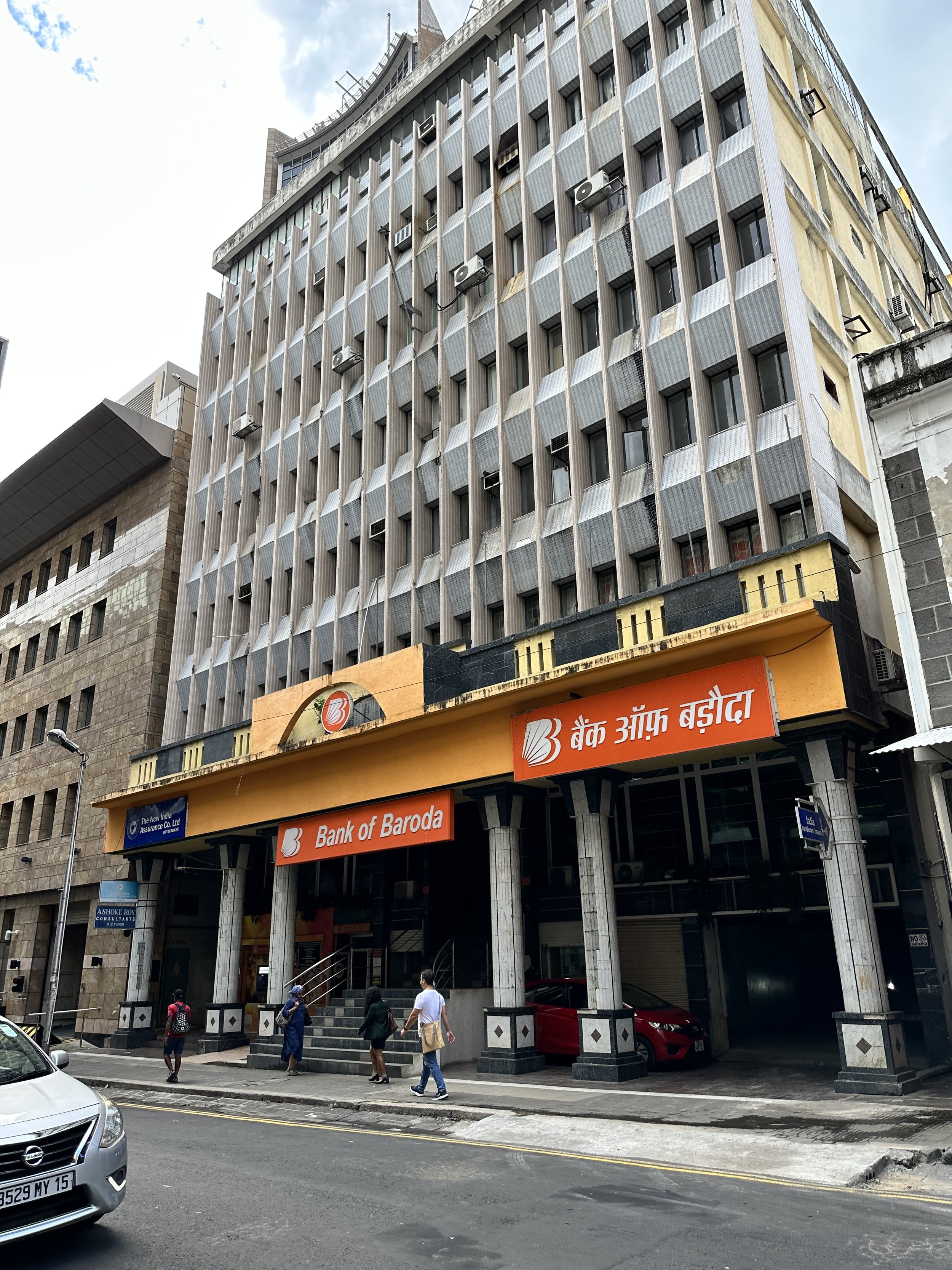 Multi-story building with a bank named 'Bank of Baroda' on the ground floor, several people walking on the sidewalk, parked cars, and an urban street scene.