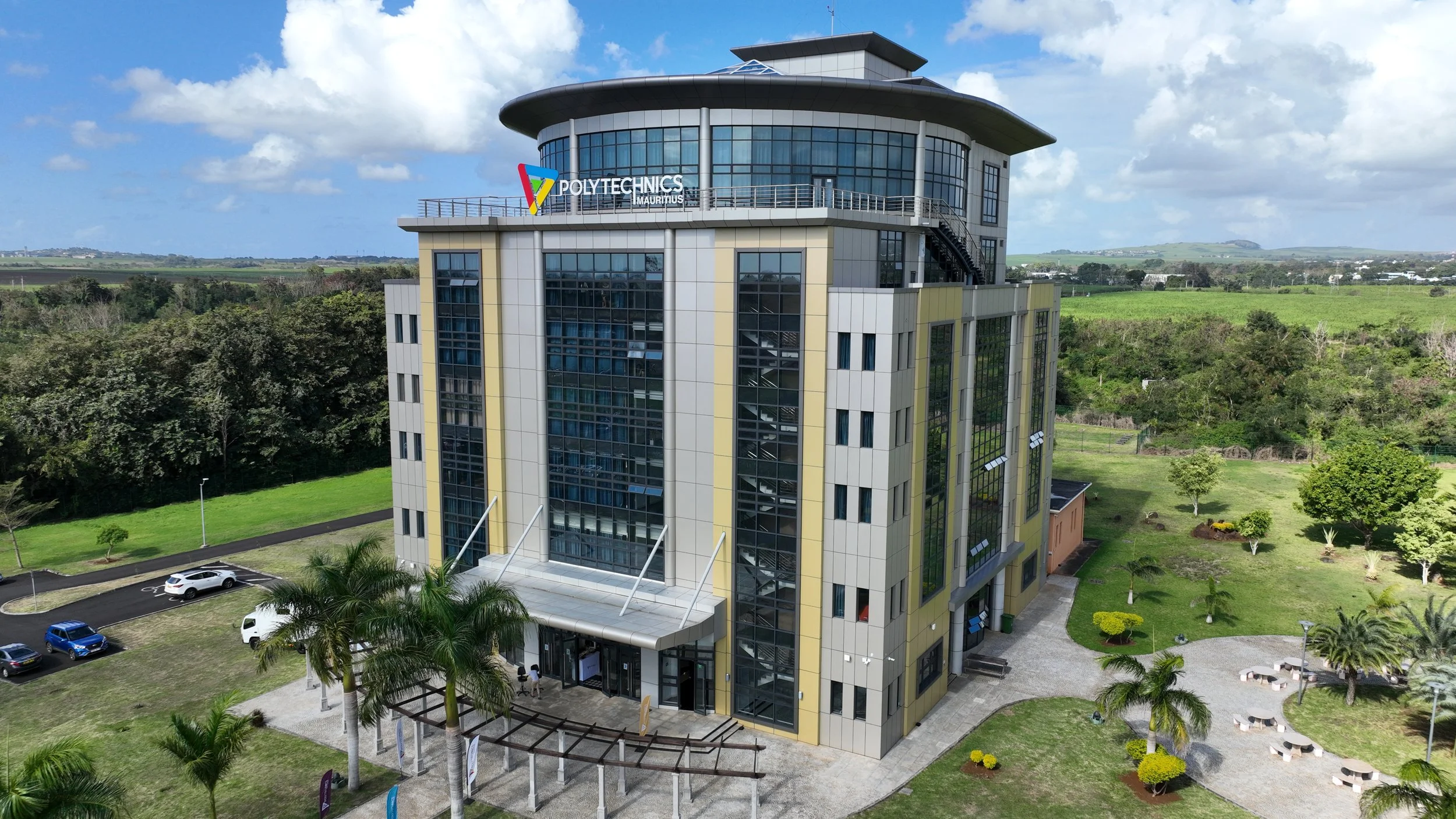 Modern multi-story building with a sign that reads 'Polytechniques Mauritius', surrounded by green lawns, trees, and parked cars, with a partly cloudy blue sky overhead.