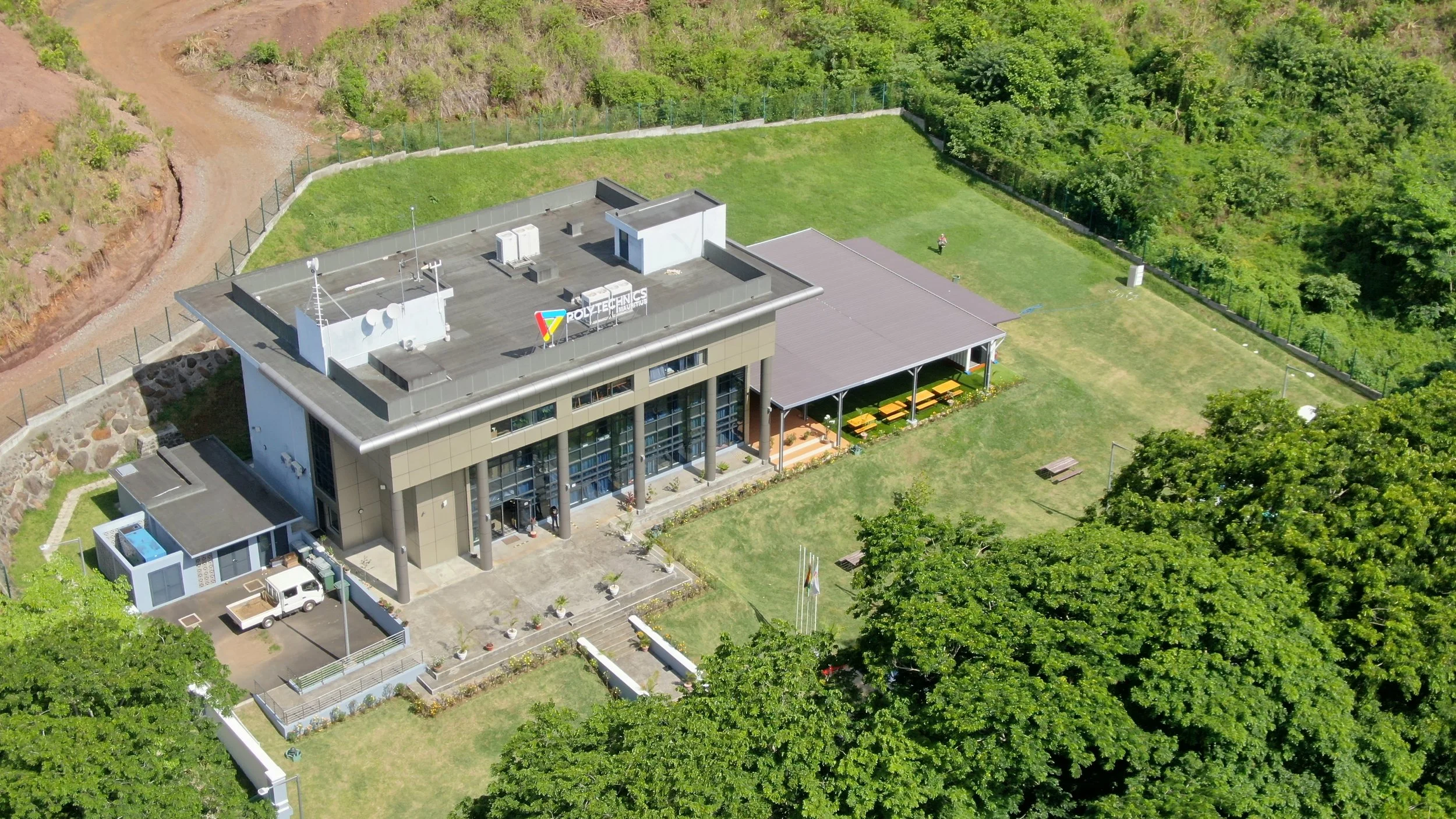 Aerial view of a modern building labeled Polytechnics, surrounded by green grass, trees, and a fenced area, with a parking space and outdoor seating area.