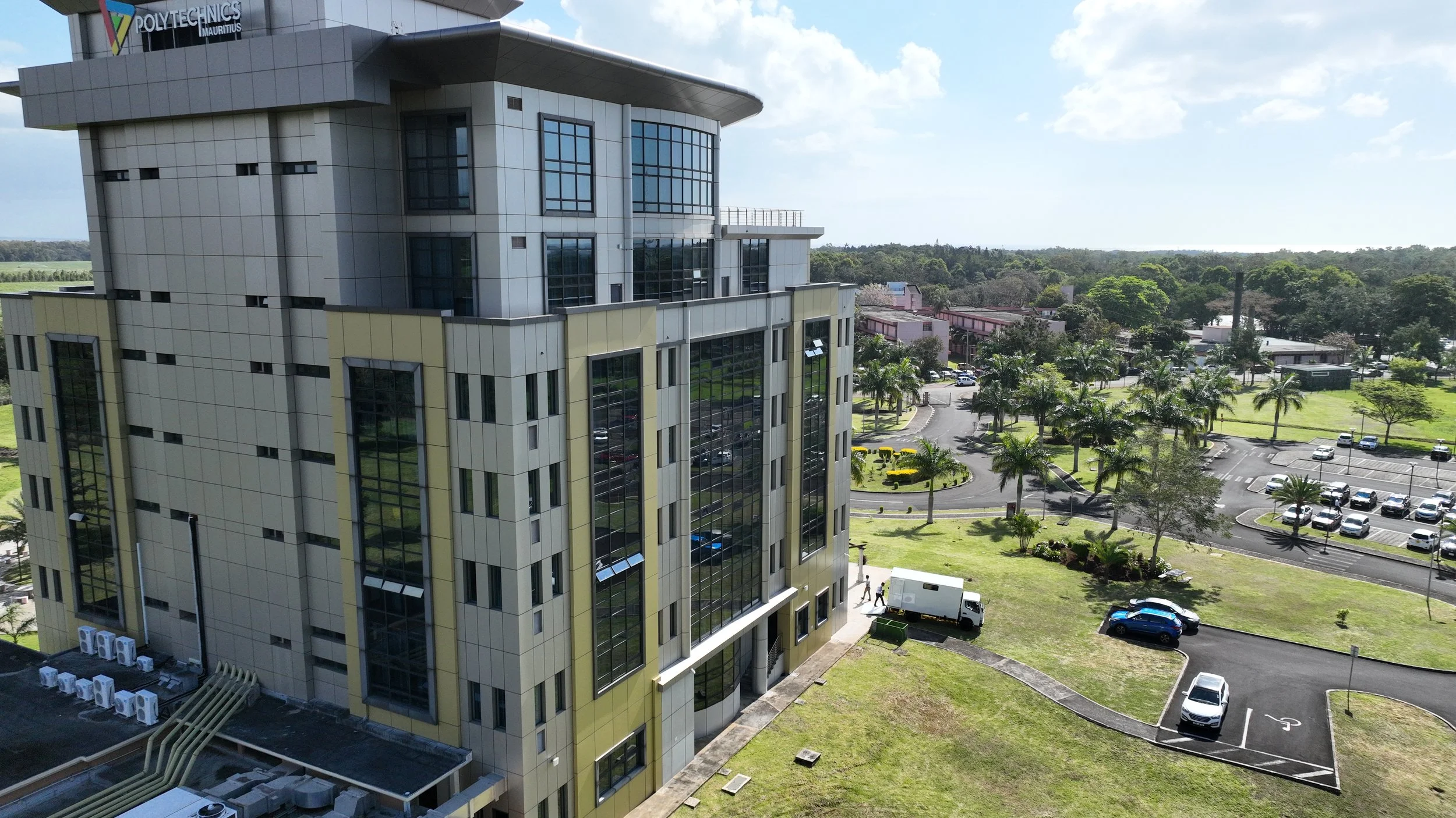 Aerial view of a modern multi-story office building with large glass windows, surrounded by parking lot, green lawns, and palm trees, under partly cloudy sky.