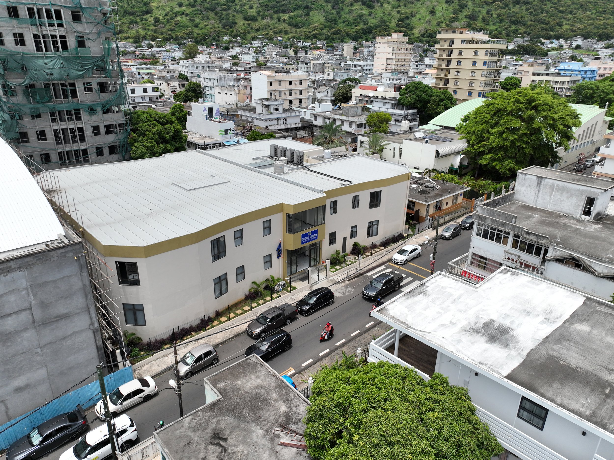 An aerial view of a cityscape featuring a white building with a curved yellow line on its roof, surrounded by other buildings, trees, and parked cars on the street.