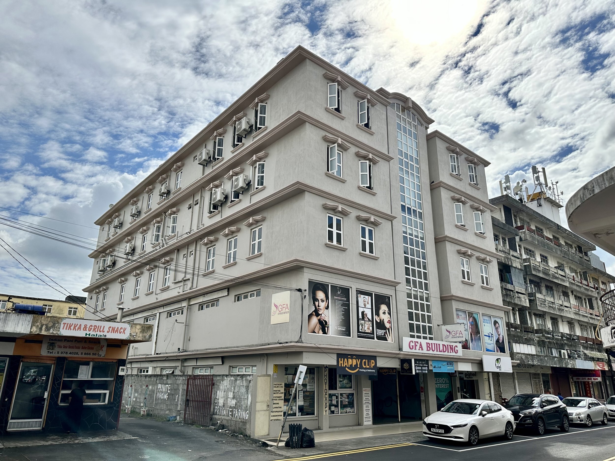 A multi-story beige commercial building with large windows and air conditioning units on the exterior. There is a sign for GFA Building, and some advertisements featuring models. The street in front has parked cars and a small food kiosk with a sign for Tikka & Grill Snack.