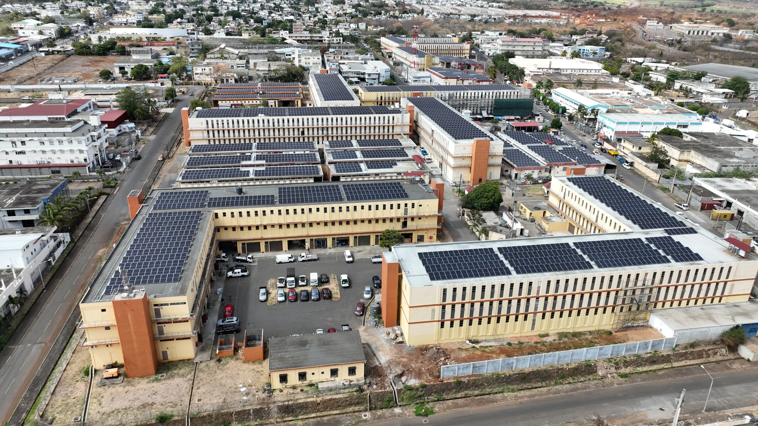Aerial view of a large complex of buildings with solar panels on the rooftops, surrounded by streets and a city landscape.
