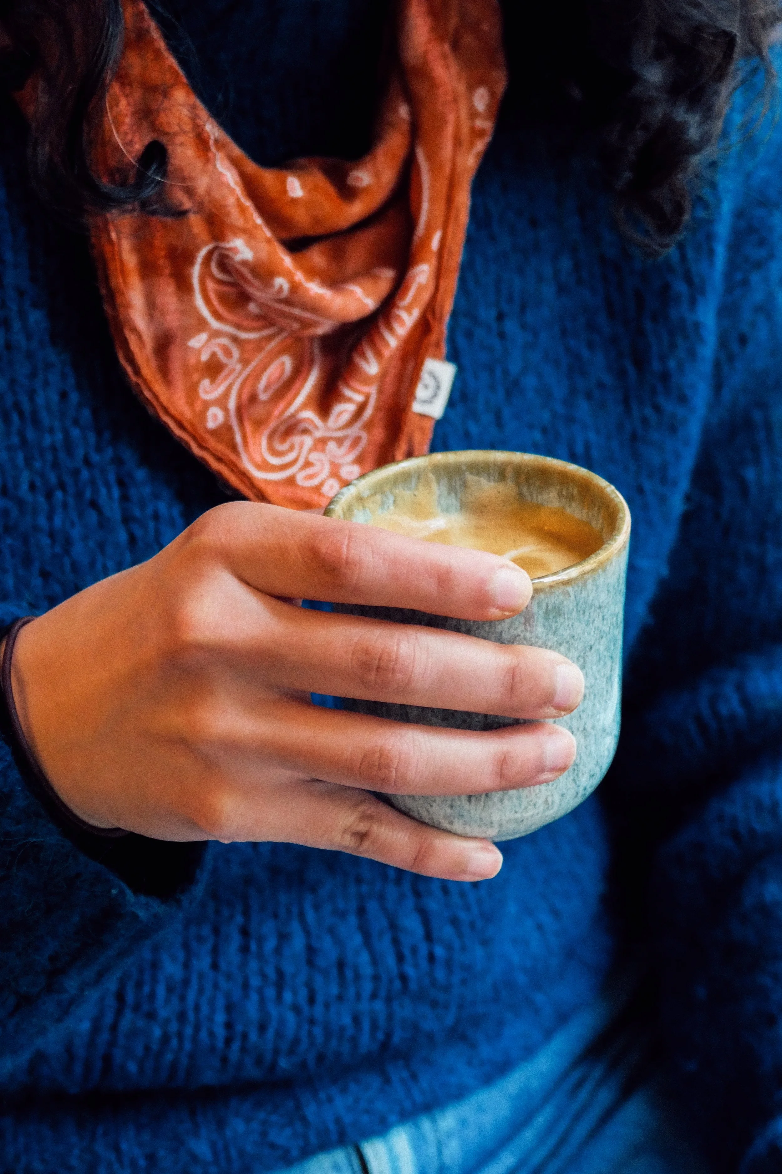 Person holding a ceramic cup of coffee, dressed in a blue sweater and an orange bandana around their neck.