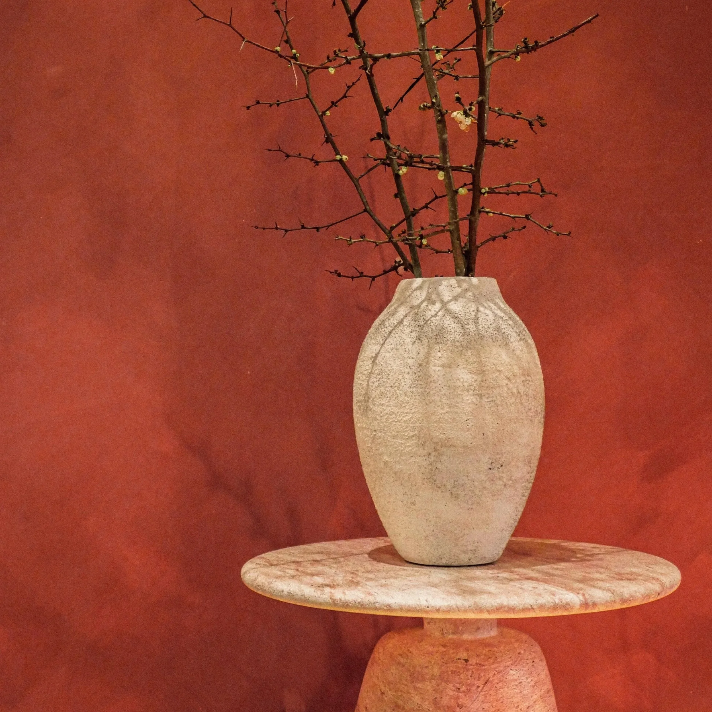 A white textured ceramic vase with a narrow neck holding bare, small, budding branches. The vase is placed on a round, stone table with a reddish wall in the background.