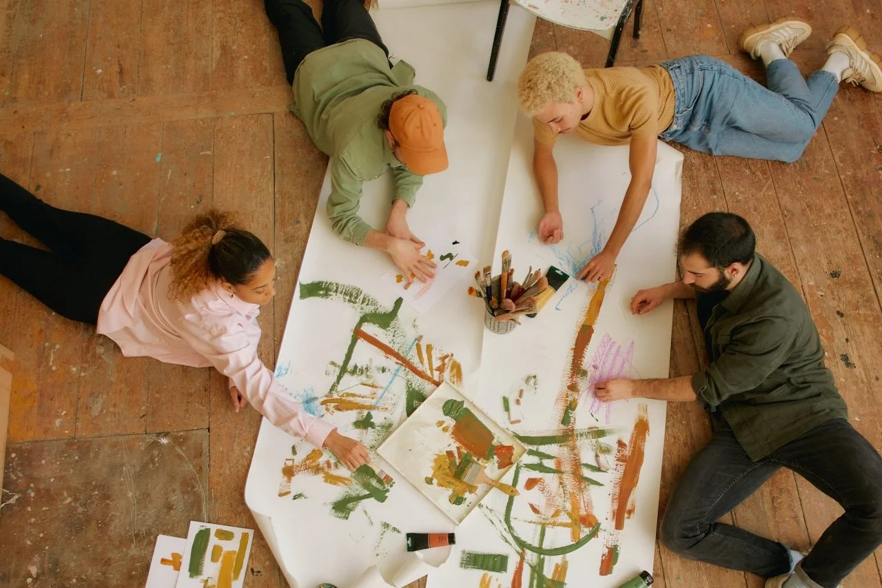 Four people lying on the floor around a large sheet of paper, painting with various colors of paint and brushes in a collaborative art activity.