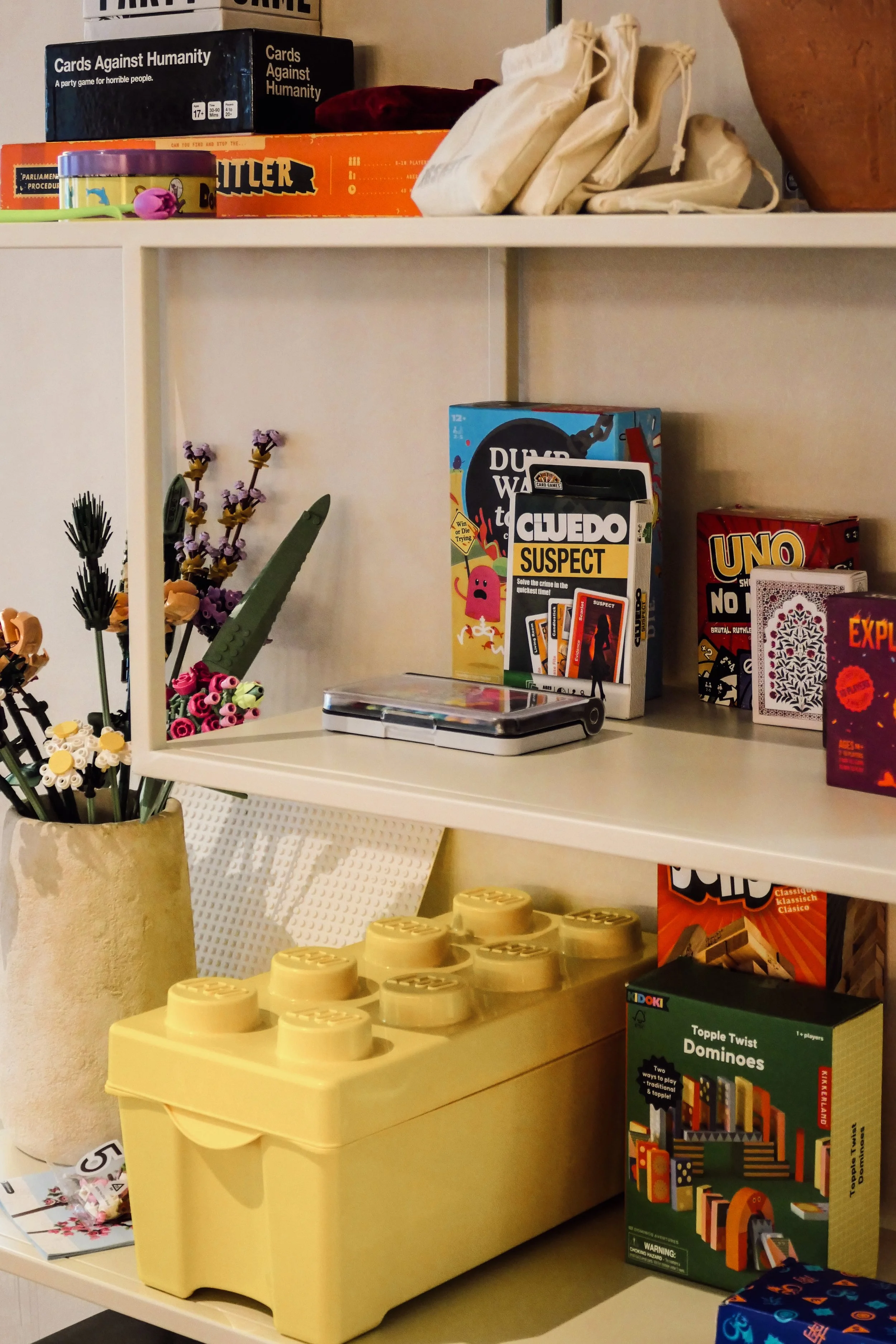 Shelf with various board games, a potted plant with artificial flowers, and a large yellow toy LEGO brick container.