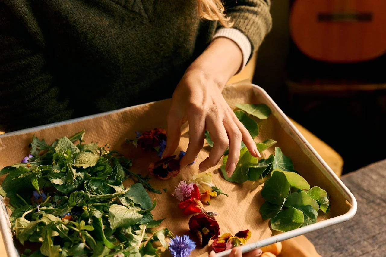 A person is arranging or selecting colorful flowers and green leaves on a tray or surface in a cozy indoor setting.