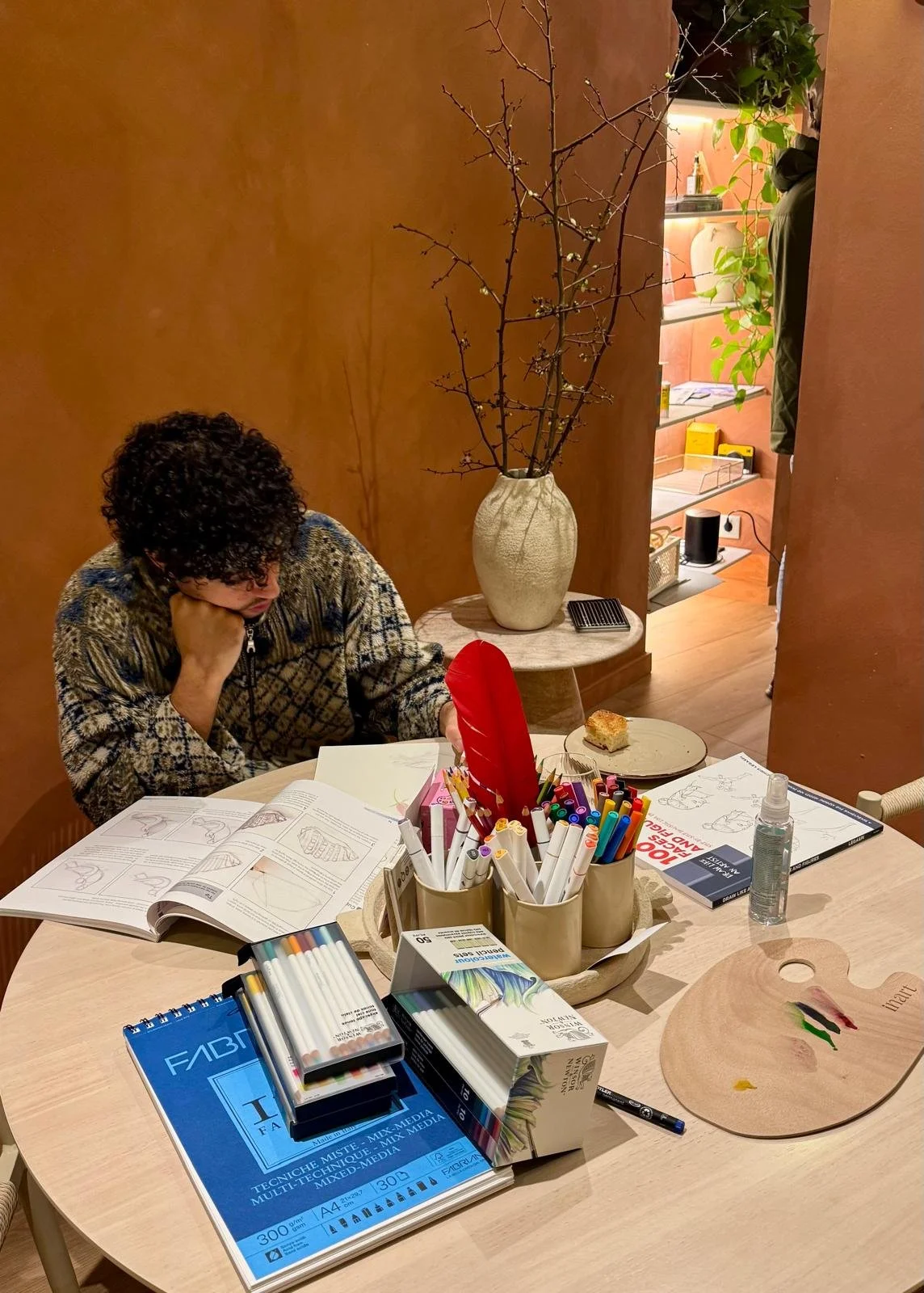 A person with curly hair sitting at a table with art supplies, open books, and a partially completed drawing on a wooden palette. The table has containers filled with markers and colored pencils, and there's a slice of cake on a plate. In the background, a large vase with bare branches and a shelf with decorative items are visible.