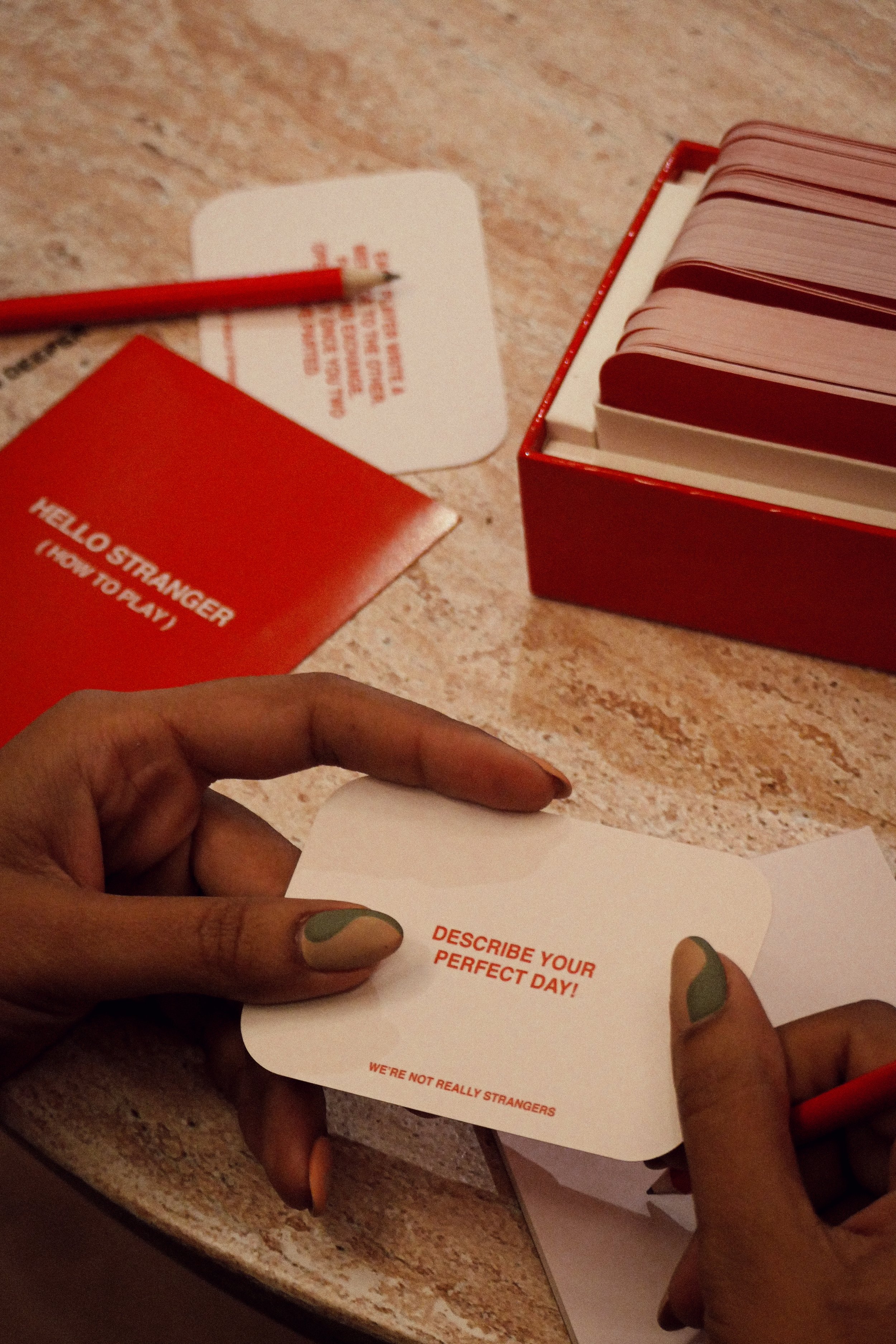 Person holding a white card with the text 'Describe your perfect day!' in red, and a red pencil. On the table are a red booklet titled 'Hello Stranger (How to Play),' a red box filled with similar cards, and a white card with red text and a list of instructions.