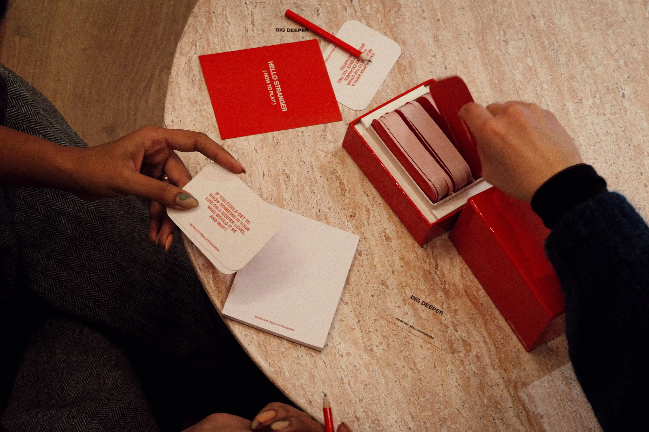 Two people sitting at a round table with a red box of cards and several cards and notes on the table. One person is holding a card, and the other person is reaching into the box.