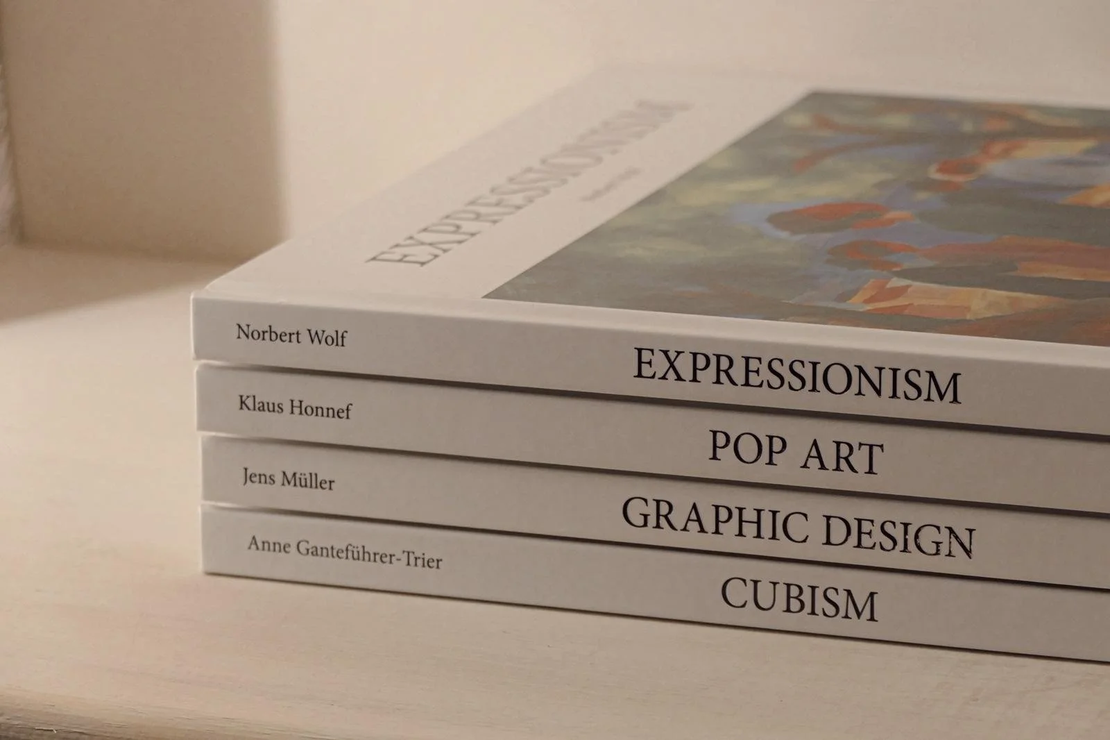 Four stacked books on art topics with white covers and black text, titled 'Expressionism, Pop Art, Graphic Design, Cubism' by different authors, on a light-colored surface with a neutral background.