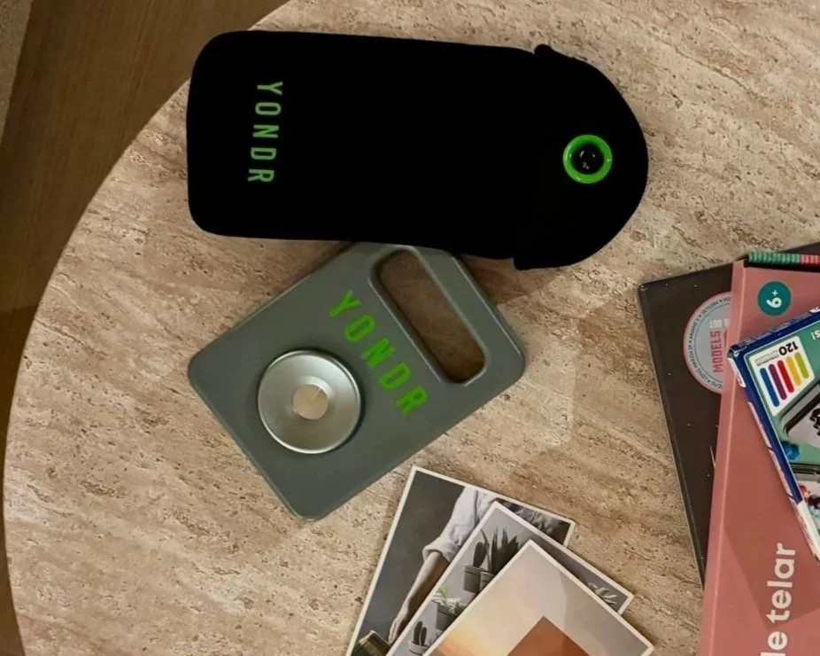 A table with a black portable charger labeled "HONOR," a gray cardholder with a circular metal ring also labeled "HONOR," some photographs, and a box of colored pencils or markers.