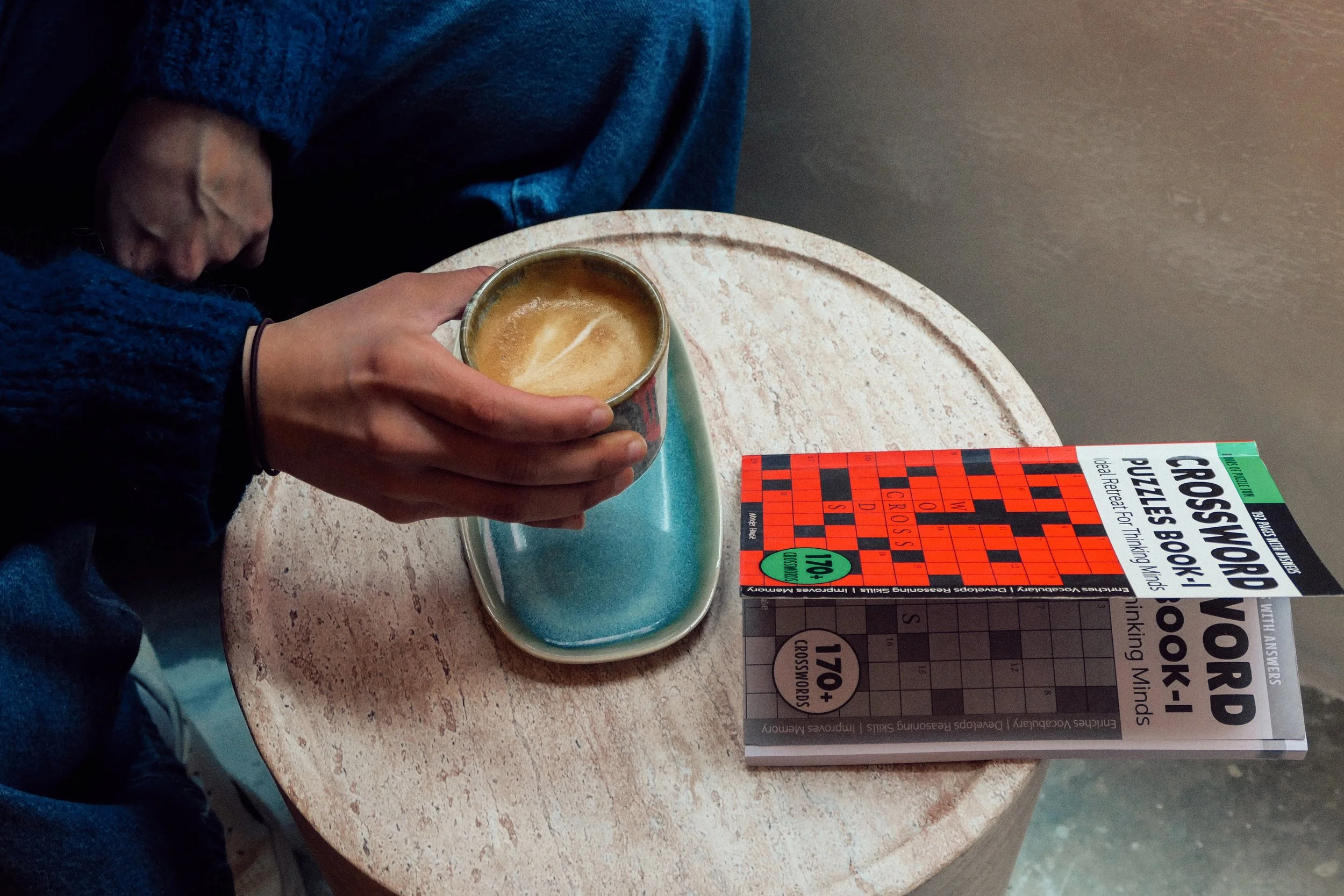 A person holding a coffee mug, sitting at a round table with a crossword puzzle book and a small tray.