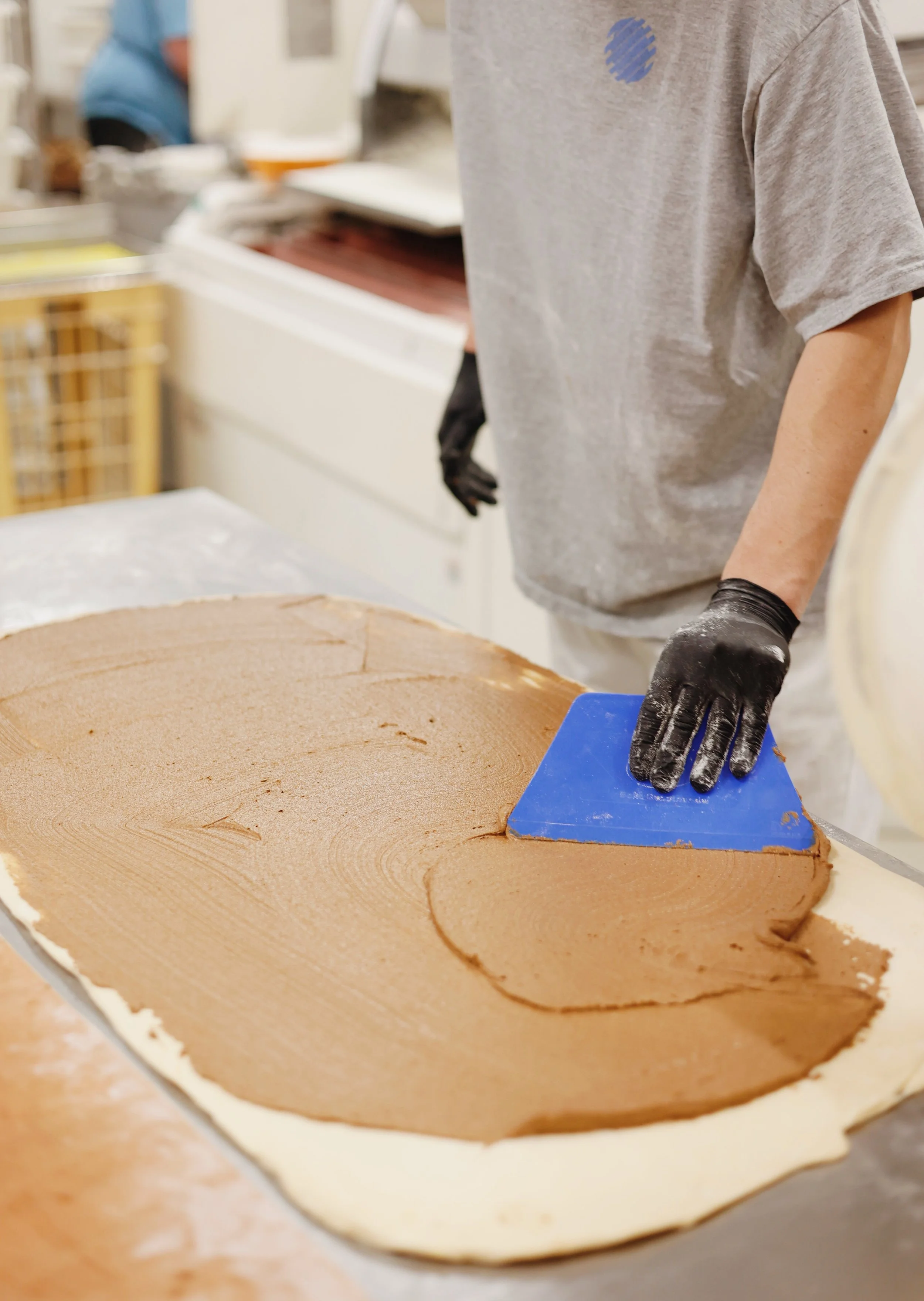 A person wearing a gray shirt and black gloves spreading chocolate filling on a large rolled-out dough sheet using a blue spatula.